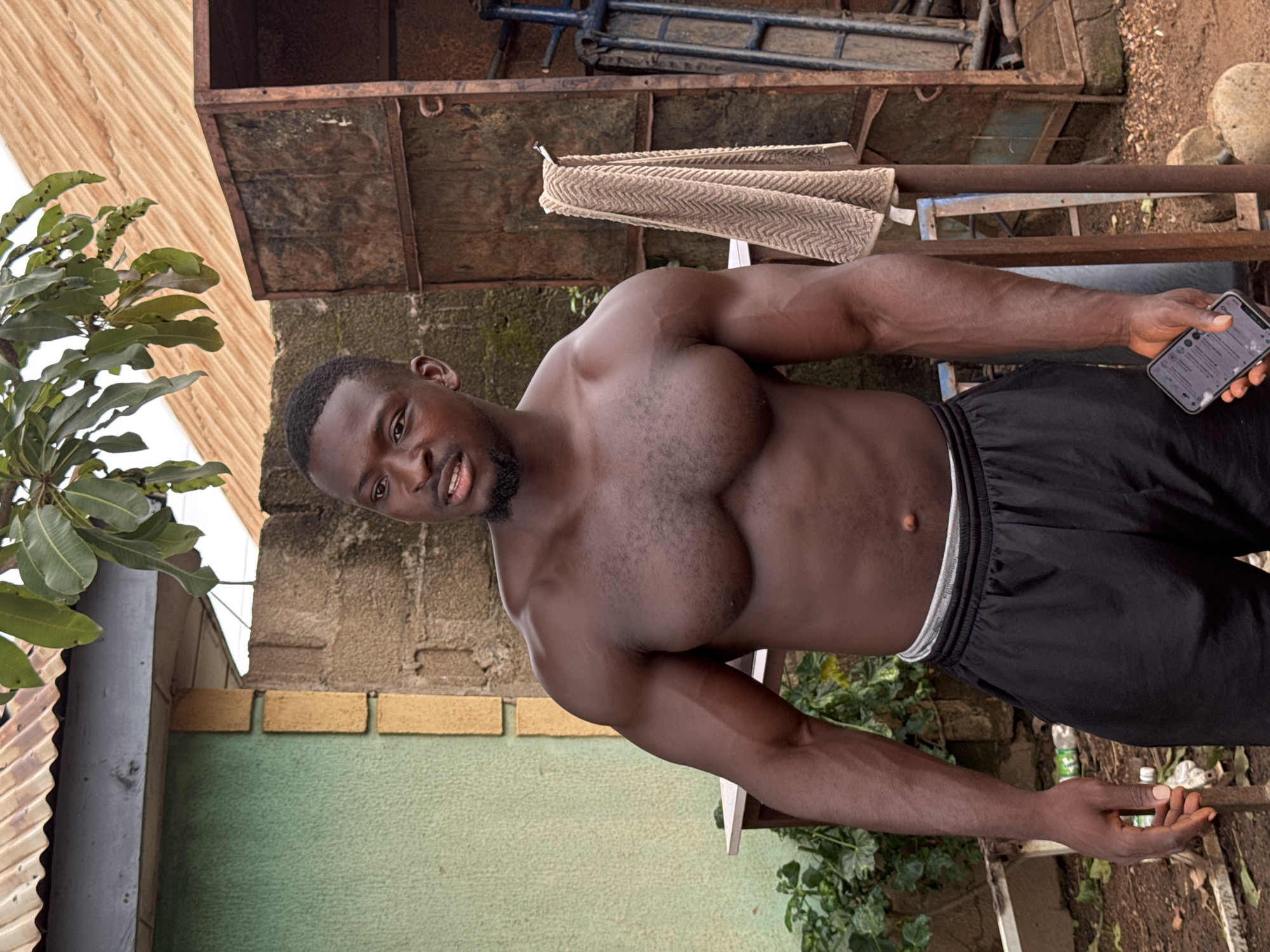 A muscular Black man, shirtless and wearing black shorts, stands outdoors in what appears to be a semi-urban or rural setting in Akwanga, Nigeria. He is looking to his left, with his mouth slightly open, and holding a smartphone in his right hand. The background features a rough, textured wall, a corrugated metal roof with a grey overhang, and some green foliage. To the right, a rusty metal structure with a towel hanging on it is visible, along with some metal framing and large stones on the ground. The overall environment suggests a somewhat unkempt outdoor area, possibly a yard or workshop.