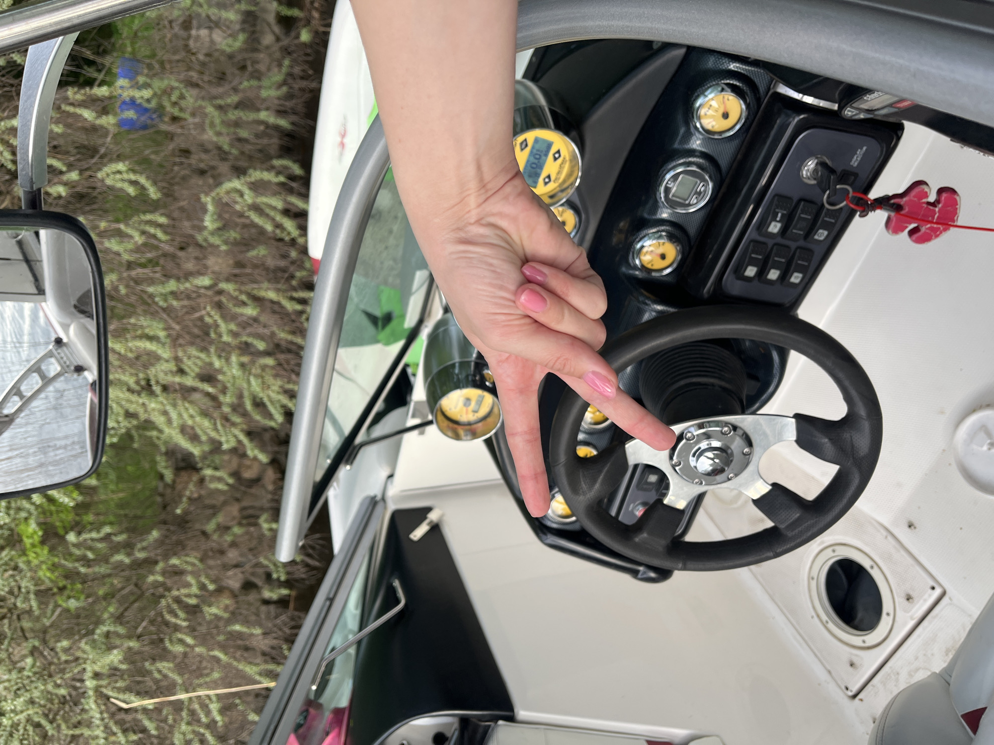 A view from inside a boat shows a hand gesturing with the index and middle fingers extended towards the steering wheel. The boat is parked near a wooded area. The location is not discernible from the image alone.