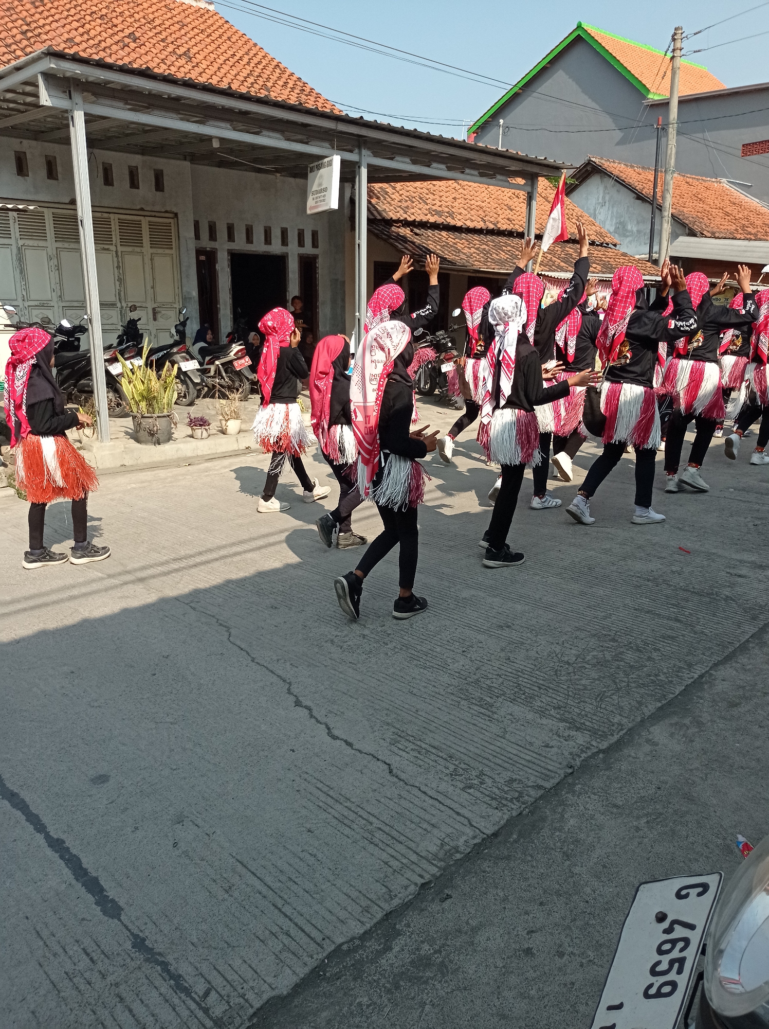 In the city of Maribaya, Indonesia, a group of approximately ten individuals, mostly young people, are participating in what appears to be a parade or performance. They are dressed in matching black tops with red and white fringe skirts, and many are wearing red headscarves. Several of the participants have their arms raised in the air. In the background, a building with an orange tiled roof is visible, as are several parked motorcycles. The ground is paved with concrete.