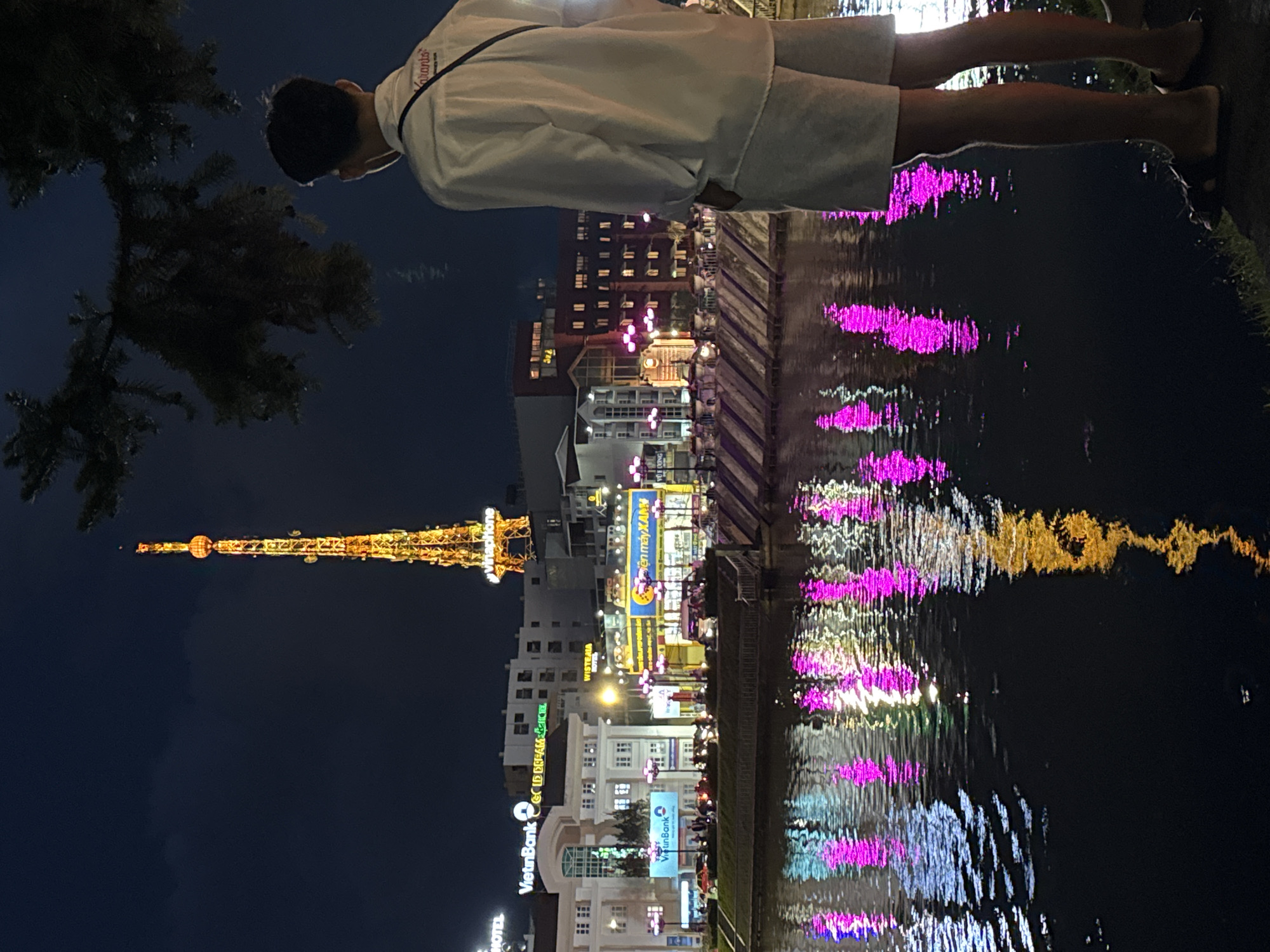 The image depicts a nighttime scene in Da Lat, Vietnam, focusing on the Xuan Huong Lake area. In the foreground, a young man stands facing away from the camera, looking out at the cityscape. He is wearing a white long-sleeved shirt and light-colored shorts, with a dark bag slung over his shoulder. The background features a prominent radio tower illuminated with yellow lights, and various buildings also lit up, casting reflections onto the dark water of the lake. The water's surface is rippled, distorting the reflections of the lights into streaks of pink, yellow, and white. The overall atmosphere is one of urban nighttime tranquility.