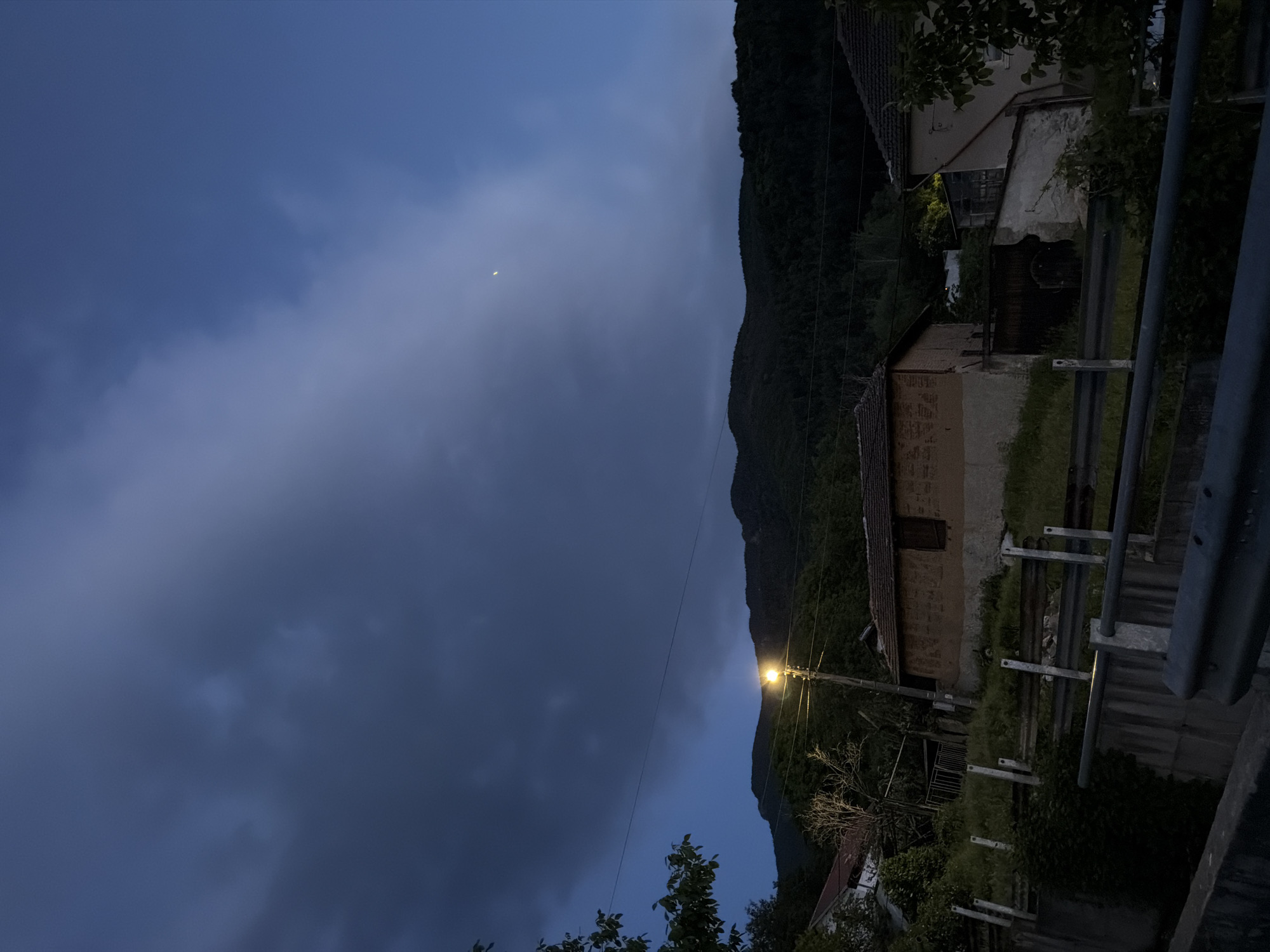 The image depicts a nighttime scene in Sapareva Banya, Bulgaria. In the foreground, there is a metallic guardrail, and beyond it, a building with a tile roof and concrete walls. A street lamp illuminates the building and a section of the nearby forest that ascends a mountain. The sky is overcast with heavy, dark clouds, and a sliver of a crescent moon is visible. Power lines stretch across the scene. The environment suggests a rural or semi-rural area on the outskirts of the town.