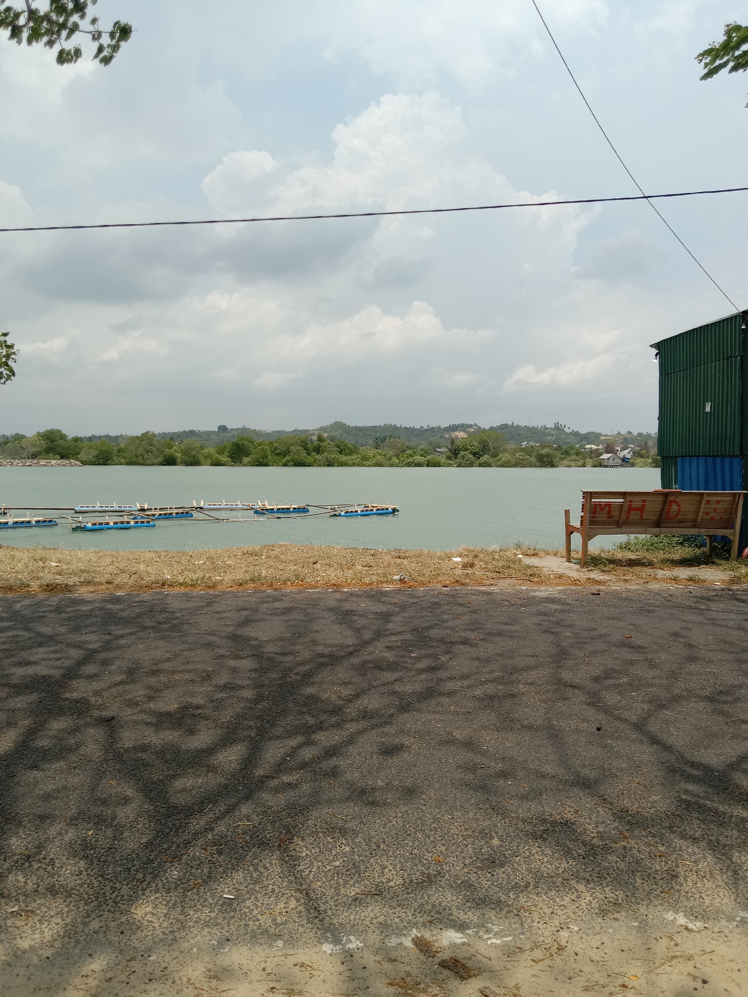 The image shows a view of a lake or river from the shoreline. A series of connected blue floats are visible on the water. On the right side of the frame, a wooden bench sits next to a green metal structure. A paved area, possibly a road, is in the foreground, covered by shadows.
While not explicitly stated, the location is likely in Lhokseumawe, Indonesia, based on the prompt. The scene lacks people and focuses primarily on the natural environment and man-made structures near the water.