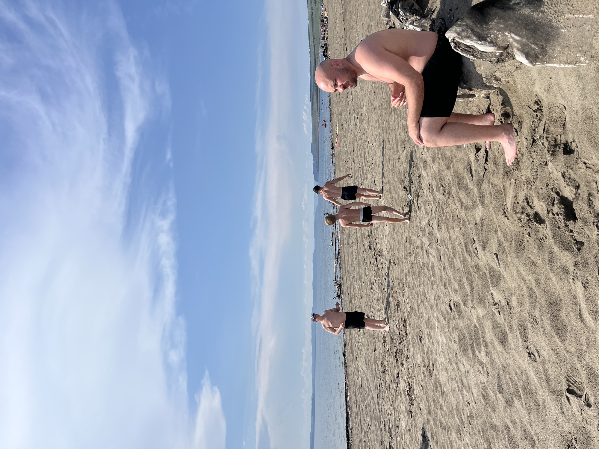 A group of four shirtless men in black swim trunks are on a sandy beach. One man in the foreground, bald and with a beard, is bent over with his hands on his knees, looking down. Behind him, three other men are standing further down the beach. The two men closest to the foreground are standing with their backs to the camera, one slightly in front of the other. The furthest man is facing away from the camera, with his left arm extended behind him. The beach is sandy with footprints visible. The sea is in the background, with a hazy shoreline and mountains in the distance. The sky is blue with wispy white clouds. The overall lighting suggests it is daytime, likely late morning or early afternoon, with sunny weather. The image appears to be taken in Krasnoturansk, Russia.