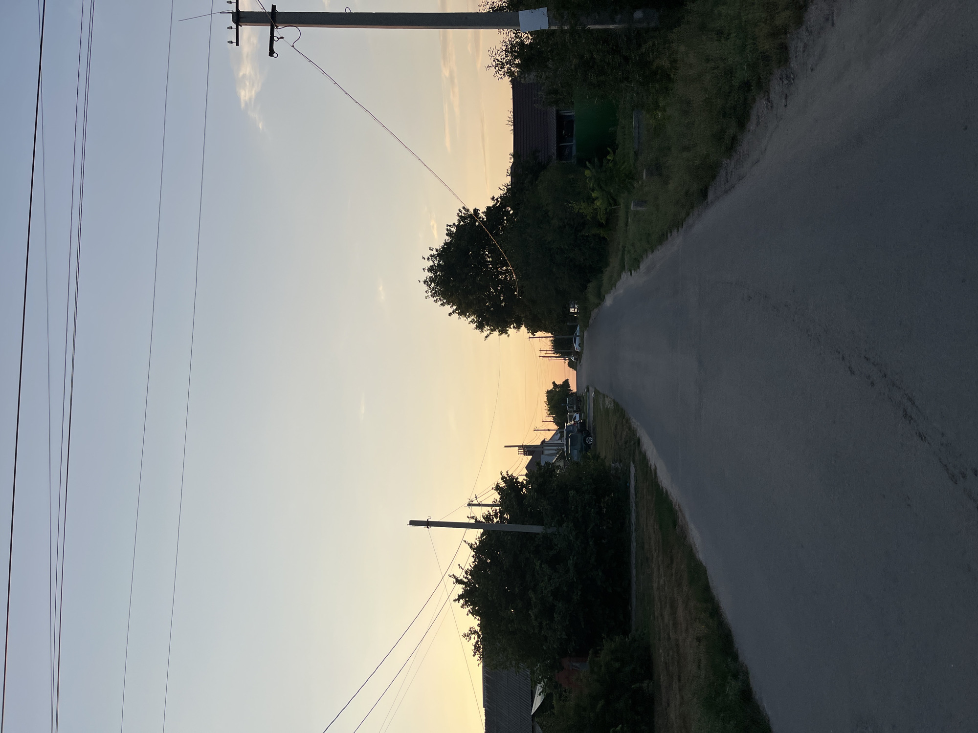 This is a photograph of a road in a rural or suburban area, possibly at dusk or dawn, with a soft, warm glow on the horizon. The road appears to be paved and is lined with trees, houses, and utility poles with overhead wires. The sky is clear with a gradient of soft blue to pale orange.
The composition is centered on the road, which recedes into the distance. On the left side of the road, there are houses with dark roofs and dense green foliage. Utility poles with multiple wires are visible, some on the left and more prominently on the right side of the road. Several cars are parked along the roadside, some partially obscured by trees and bushes. The overall impression is one of a quiet, possibly residential, street.
No specific landmarks or identifying features are visible that would allow for pinpointing a precise location within Saratov, Russia. The scene is generic enough to be found in many similar rural or suburban settings.