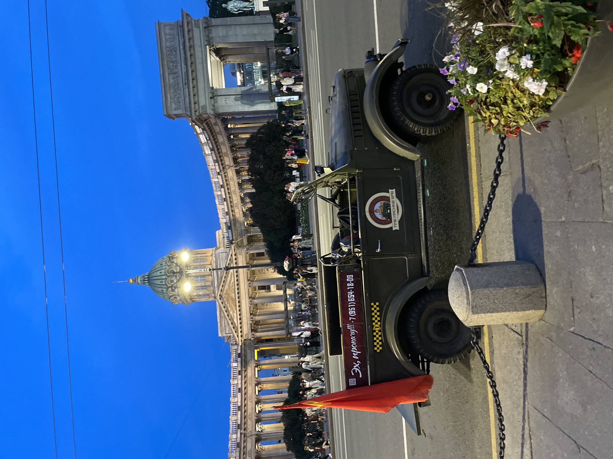 The image depicts a street scene at dusk, with the Kazan Cathedral in St. Petersburg as a prominent backdrop. In the foreground, a vintage olive-green military-style vehicle, possibly a GAZ-69, is parked on the street. A red flag is attached to the rear of the vehicle, and text, including what appears to be a phone number, is visible on its side. The vehicle has a "TIME MACHINE SPB" logo on its door.
A crowd of people are walking along the sidewalk in front of the cathedral, which is illuminated and features a large dome and colonnades. The sky is a deep blue, transitioning from twilight to night. A planter with flowers and foliage is visible in the lower right corner of the image, and a decorative stone bollard with a chain barrier separates the sidewalk from the street. The overall impression is of a tourist area, with the vintage vehicle likely serving as a themed attraction.