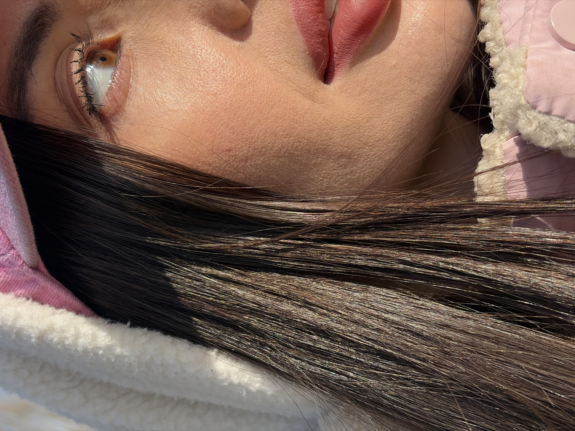 Close-up portrait of a person with dark brown hair, light skin, and light brown eyes. The person is wearing a pink jacket with a white fluffy collar. The background is out of focus. The location cannot be determined based on the image.