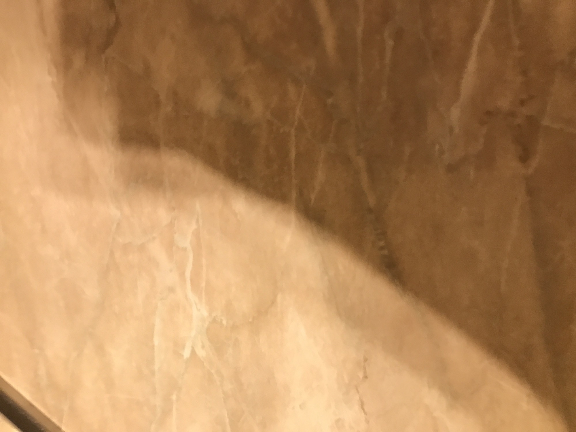 The image shows a close-up of a wall covered in light brown marble with darker brown veins. A shadow falls diagonally across the surface, obscuring some of the marble pattern. There is a dark horizontal line visible at the lower-left edge of the frame. The image contains no discernible people, subjects, or objects aside from the marble wall and shadow. No location information is discernible from the image.