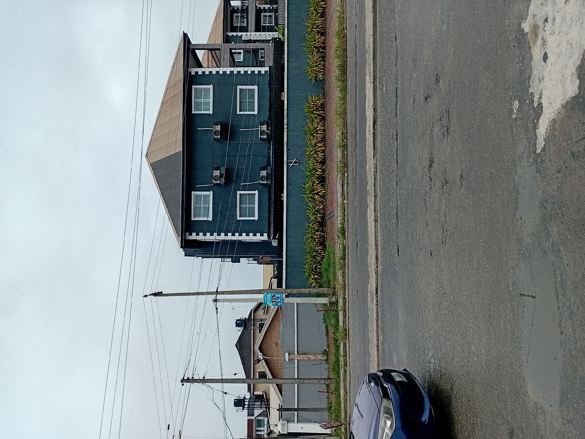 The image shows a street scene in an urban environment under an overcast sky. A dark blue car is partially visible in the lower left corner, its front end facing left. The road surface is wet and appears to have puddles. In the mid-ground, a dark blue building with white trim and multiple windows is prominent. Several air conditioning units are mounted on the exterior wall of this building. A low, teal-colored wall runs in front of the building, with manicured bushes and some dry grass growing along its base. Beyond the wall, other buildings and structures are visible, including a house with a brown roof and water tanks on top, and another multi-story building. Electrical poles and wires are scattered throughout the scene, with a poster attached to one of the poles. The overall atmosphere suggests a damp or post-rain environment.