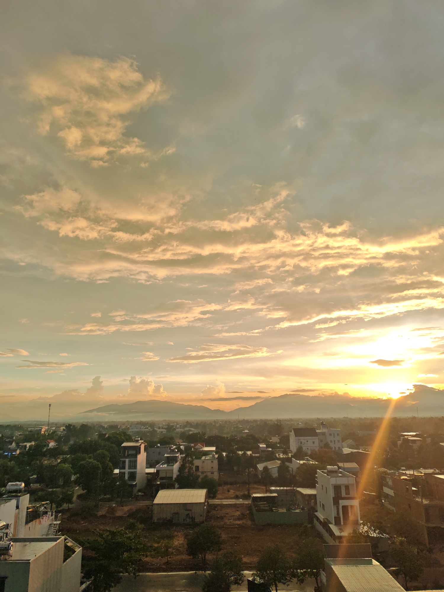 This is a high-angle, vertical shot of a city at sunrise. The sky is filled with dramatic clouds tinged with orange and yellow from the rising sun, which is positioned on the right side of the frame. The sun's rays are creating a lens flare effect that extends diagonally across the image. Below the sky, a cityscape stretches out with low-rise buildings, many of them appearing to be residential or commercial properties. Trees are scattered throughout the urban landscape, and in the distance, mountains form a hazy backdrop. The scene suggests a developing urban area, possibly on the outskirts of a larger city, given the mix of construction and undeveloped land visible. The overall atmosphere is serene, characteristic of an early morning in a warm climate. The specific location of Đà Nẵng, Vietnam, is not visually identifiable from the image alone, but the architectural style and vegetation are consistent with tropical Southeast Asian urban environments. There are no people or specific actions depicted in the image; it is a still landscape captured at dawn.