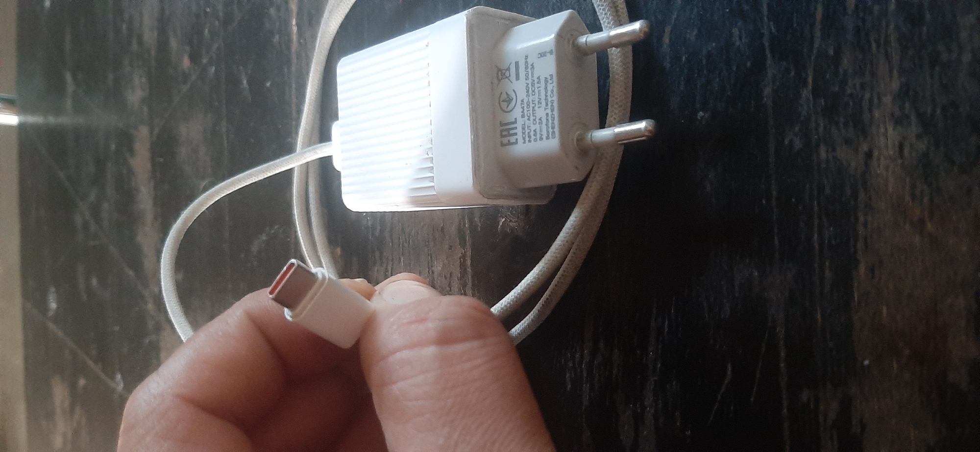 This is a close-up, high-angle, eye-level shot of a white USB-C charger and its cable, resting on a dark, textured surface, likely a wooden table. The charger is a compact, rectangular block with a ridged pattern on one side and a European-style plug on the other. Visible text on the charger indicates it is a Borofone Technology product, model BAATA, designed for AC 100-240V input, with multiple output options including 9V 2A and 12V 1.5A. The cable is white and braided, with a USB-C connector on one end, which is being held by a person's hand. The hand is slightly blurred, suggesting movement or shallow depth of field. The lighting appears to be artificial, casting subtle shadows and highlighting the textures of the charger and the surface it rests on. The overall impression is one of a functional electronic accessory, possibly being prepared for use or examination, set against a plain, everyday backdrop. There are no clear indicators of time of day, weather, or specific emotions. The location context provided suggests Yessentukskaya, Russia, but this cannot be visually confirmed from the image alone.