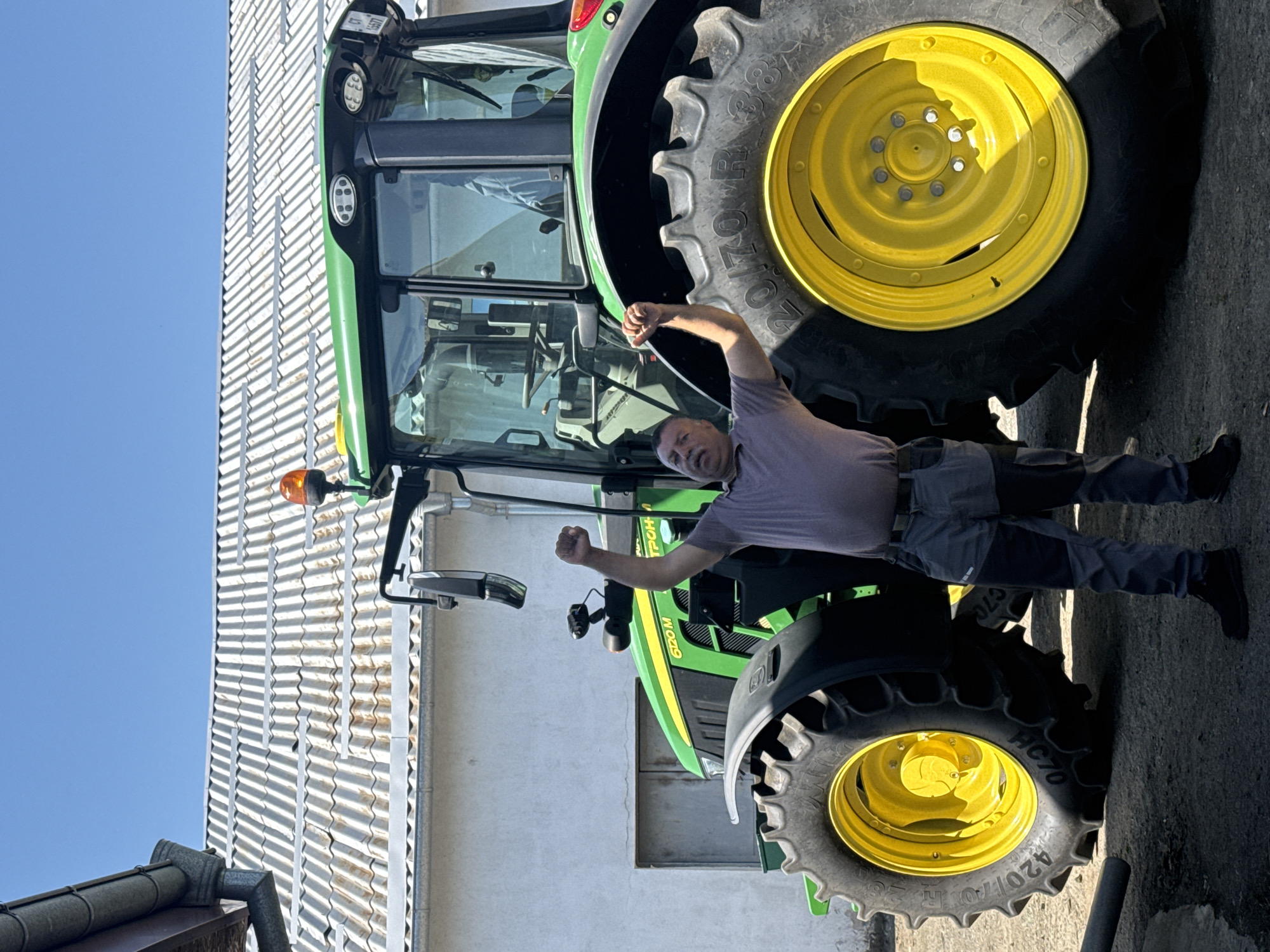 The image shows a man standing next to a John Deere tractor. The tractor has large yellow wheels and a green body. The man is wearing a grey t-shirt and grey pants. He is standing with his arms raised in a celebratory gesture. The location appears to be outdoors, possibly in a rural area or on a farm, next to a corrugated metal building. The sky is blue. There is no confirmation that the location is Sarnevo, Bulgaria based on the visual information provided.