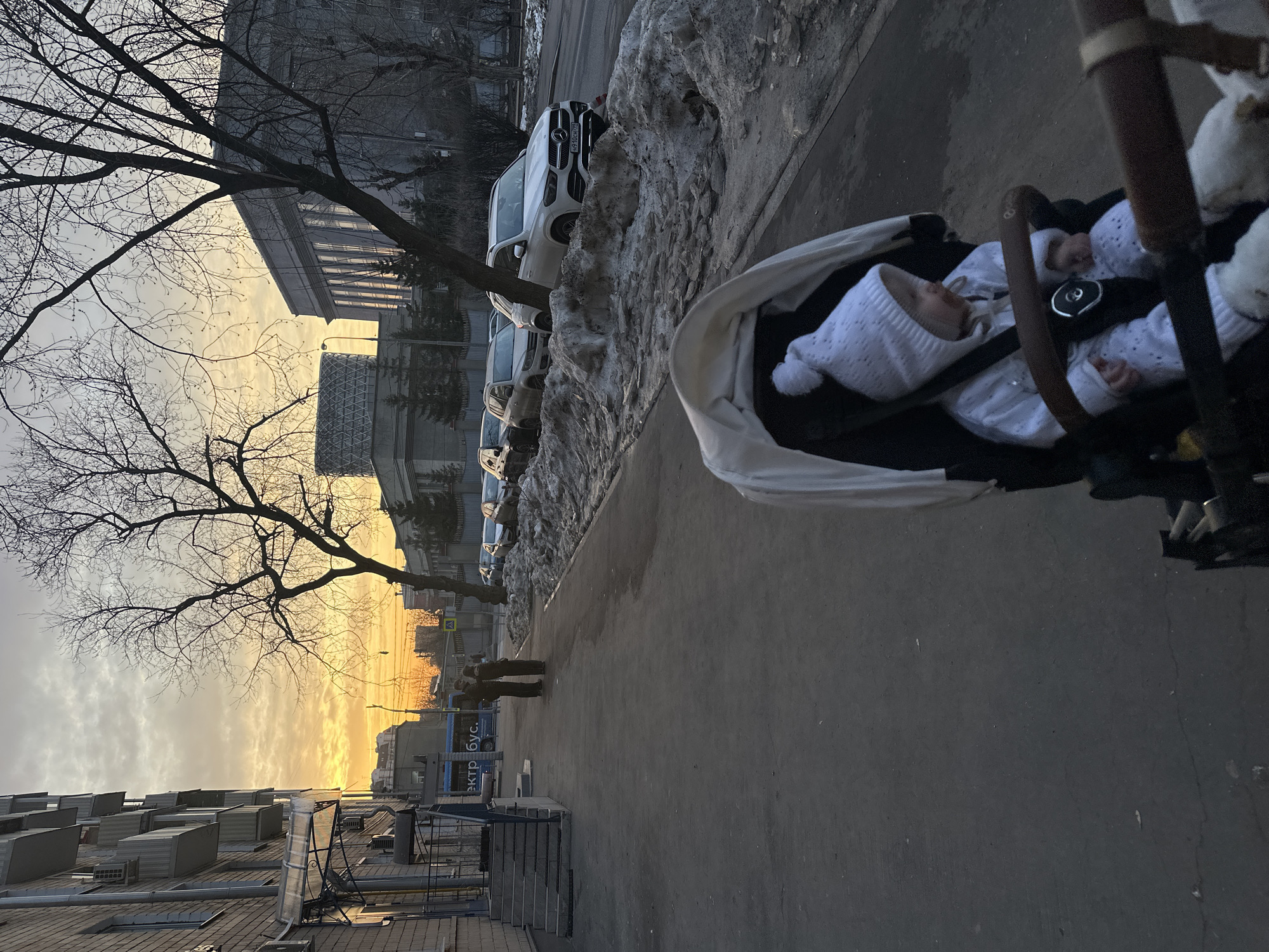 The image depicts an urban street scene in Moscow, Russia, at sunset or sunrise, characterized by a golden-orange sky. In the immediate foreground, a baby dressed in white knitwear, including a hat with a pompom, is seated in a dark stroller with a light canopy, facing slightly right. The stroller's wooden handlebar is visible. To the left, a paved sidewalk extends, where two adult figures stand facing each other in the mid-distance. To the right of the sidewalk, a street features several parked vehicles, including a white Mercedes-Benz GLE SUV with Russian license plate K910OC799. Piles of dirty, melting snow and ice line the curb between the sidewalk and the street. On the left, a multi-story brick building with balconies and air conditioning units borders the sidewalk. In the background, silhouetted bare trees partially obscure a large, grey, classical-style building with tall windows and a modern cylindrical building with a geometric grid pattern on its upper portion. A blue city bus with partial Cyrillic text "ЭКТР БУС" is also visible in the background.