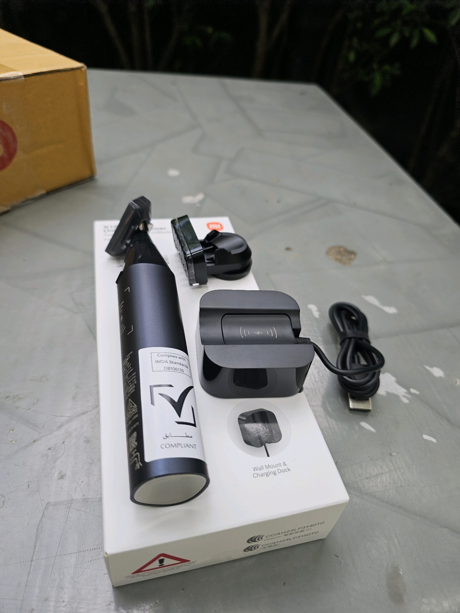 A table is outdoors. On the table are various items including a cardboard box, a black device with a trimming head, a black charging dock with a USB cable, and a black mounting bracket. The table surface is gray and appears to be made of concrete or stone. In the background, there are dark shadows and some out-of-focus green foliage, suggesting a natural or park-like setting. The lighting indicates it is daytime, possibly overcast, with shadows cast by the objects.