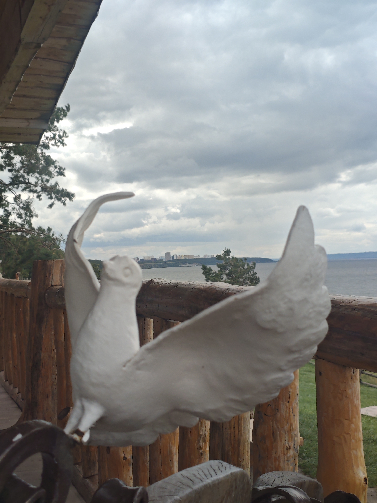 The image shows a white statue of a bird with its wings spread, positioned on a wooden railing overlooking a body of water. In the background, a city skyline with buildings is visible across the water, under a cloudy sky. A wooden roof and railing are present in the foreground. The scene appears to be outdoors, possibly in a park or recreational area, with a view of the Volga River in the city of Arkhangelskoye, Russia. There are no people visible in the image.