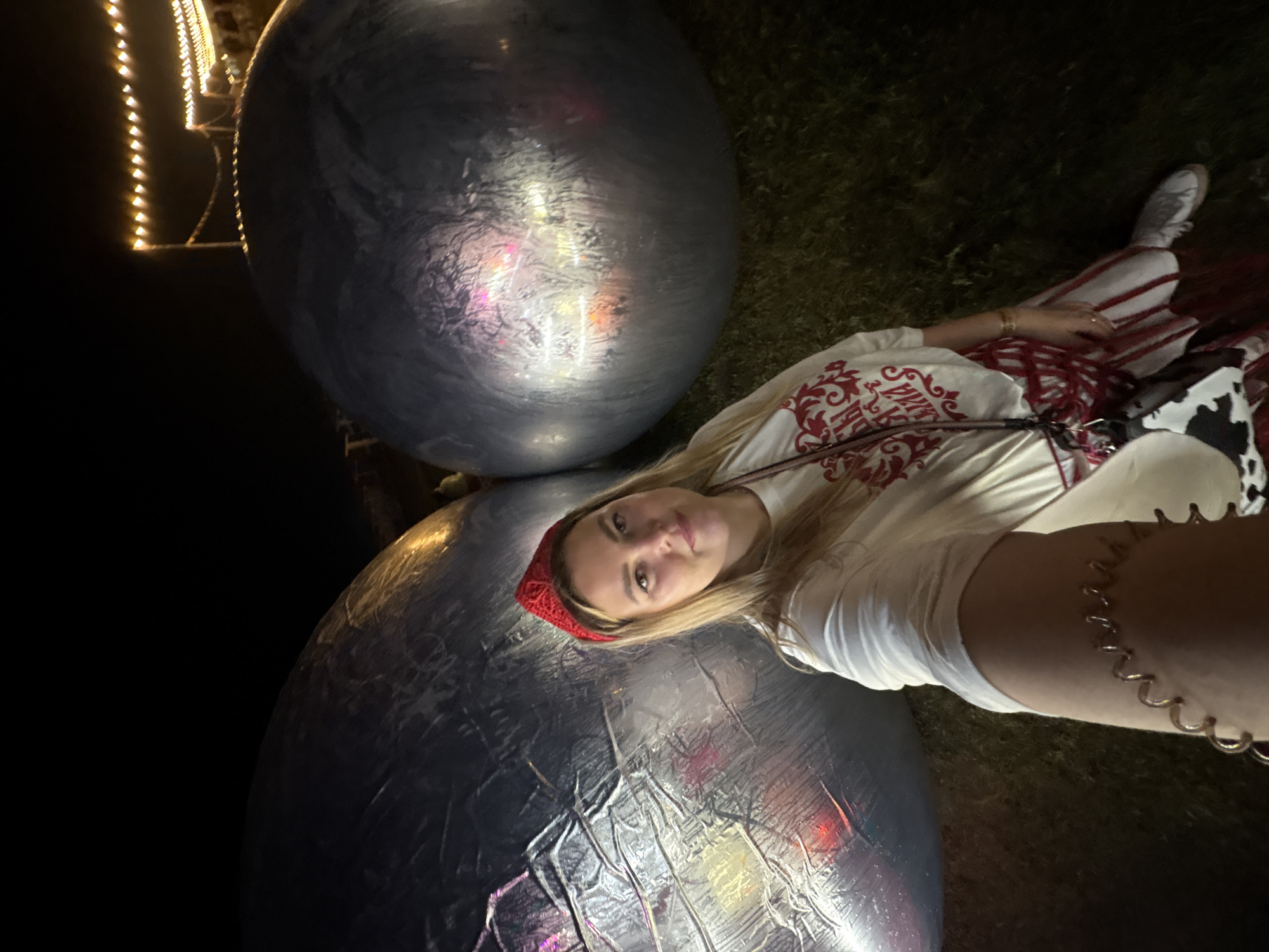 This is a selfie taken by a young woman lying down next to two large, spherical, metallic-looking objects. She is wearing a white t-shirt with red ornamental designs and a red headband. She has blonde hair and is looking at the camera with a slight smile. Her outfit also includes a red and white striped skirt and white sneakers. She is also wearing a gold bracelet on her left arm, which appears to be coiled like a spring. In the background, there are some string lights with yellow bulbs, suggesting it might be nighttime or dusk. The ground appears to be covered in grass. The large spheres are reflective and appear to be illuminated by the surrounding lights. The overall mood of the image is casual and playful.