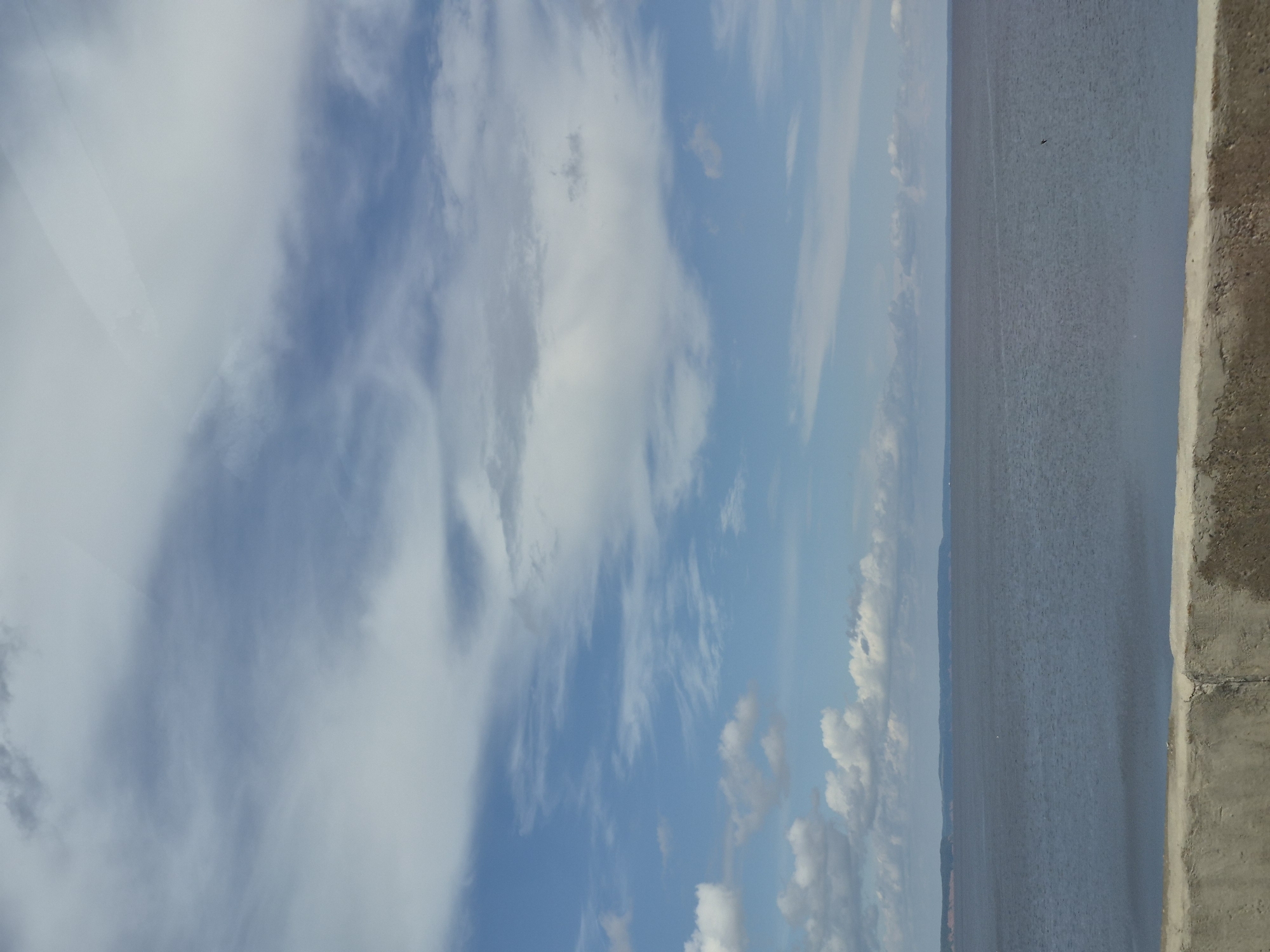 A wide, serene body of water stretches horizontally across the lower half of the image, meeting a distant, hazy shoreline. The water is a deep, muted blue, with subtle ripples indicating a gentle breeze. A rough, concrete barrier runs along the bottom edge of the frame, its texture and color suggesting it's a weathered embankment. The sky above is a vast expanse of blue, streaked with dynamic formations of white and gray clouds. Some clouds are wispy and diffused, while others are more dense and textured. The overall impression is one of natural tranquility under a partly cloudy sky, likely a scenic overlook in Chaikovsky, Russia, perhaps along the Kama River.