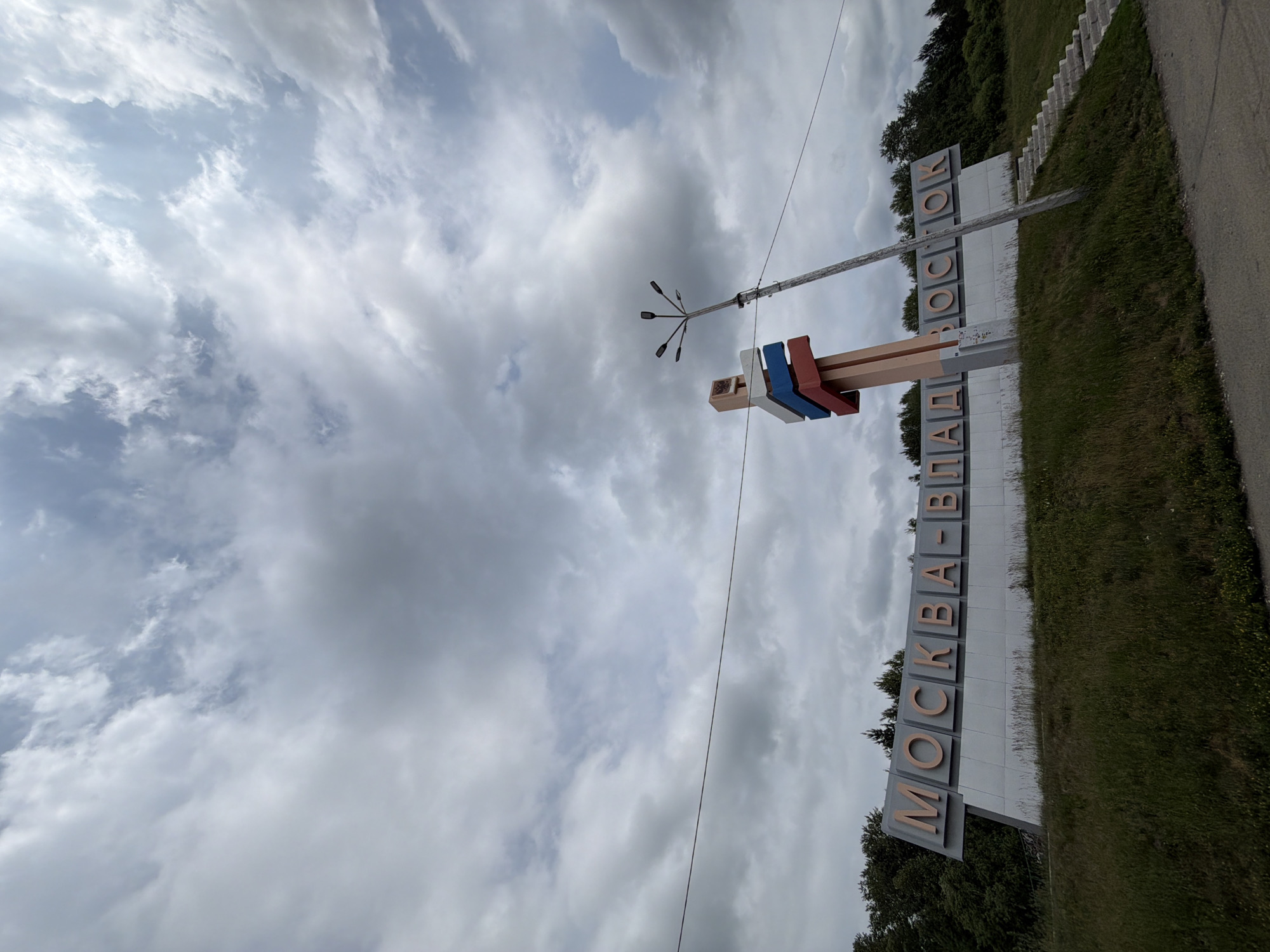 An eye-level, wide-angle shot captures a large, gray, angled sign with orange letters reading "МОСКВА-ВЛАДИВОСТОК" (Moscow-Vladivostok). The sign is situated on a grassy hill, with trees forming a dark backdrop behind it. To the right of the sign, a tall, weathered utility pole with multiple lamp fixtures extends upwards, leaning slightly to the right. A concrete staircase with several steps ascends the grassy hill to the right of the sign. In the foreground, a gray asphalt road runs along the bottom of the frame. The sky above is filled with dramatic, gray and white clouds. The scene appears to be an outdoor roadside landmark.
