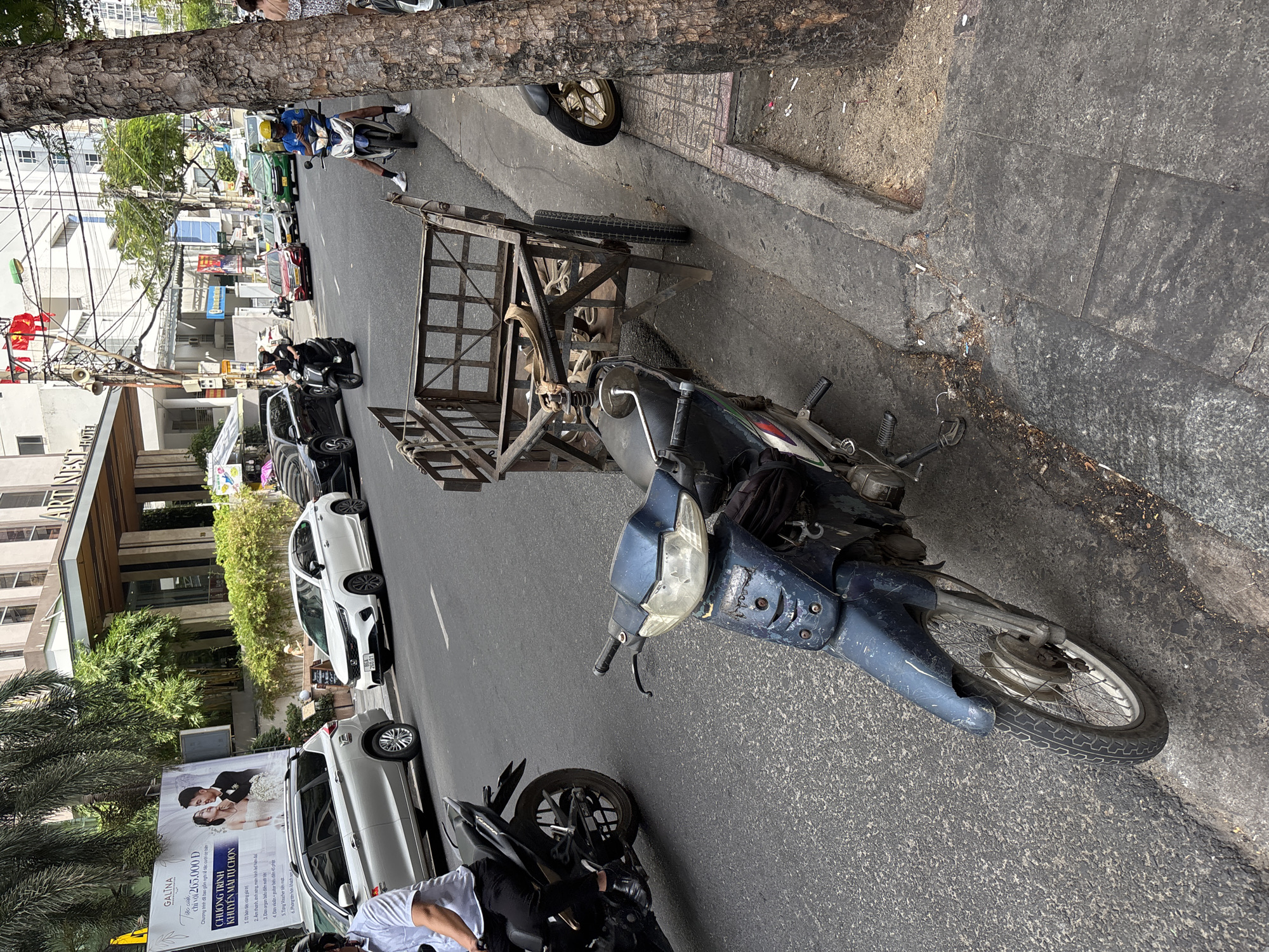 In Nha Trang Ward, Vietnam, a street scene unfolds with a focus on the right side of the frame, featuring a heavily weathered, dark blue, scooter parked by a sidewalk. The scooter is attached to a rustic metal cart with two wheels and a wooden frame, suggesting it's used for transport. To the right of the scooter, a large tree trunk with textured bark stands prominently. The sidewalk itself is paved and shows signs of wear, with a small patch of dirt and some discarded cigarette butts visible. Beyond the immediate foreground, the street is visible with other vehicles, including a white car and several motorcycles, indicating typical urban traffic. Buildings line the street in the background, one with a prominent "ARTINEST HOTEL" sign. A large billboard promoting a wedding package is also visible on the left side of the street. The overall impression is of a bustling city environment with functional, everyday scenes.