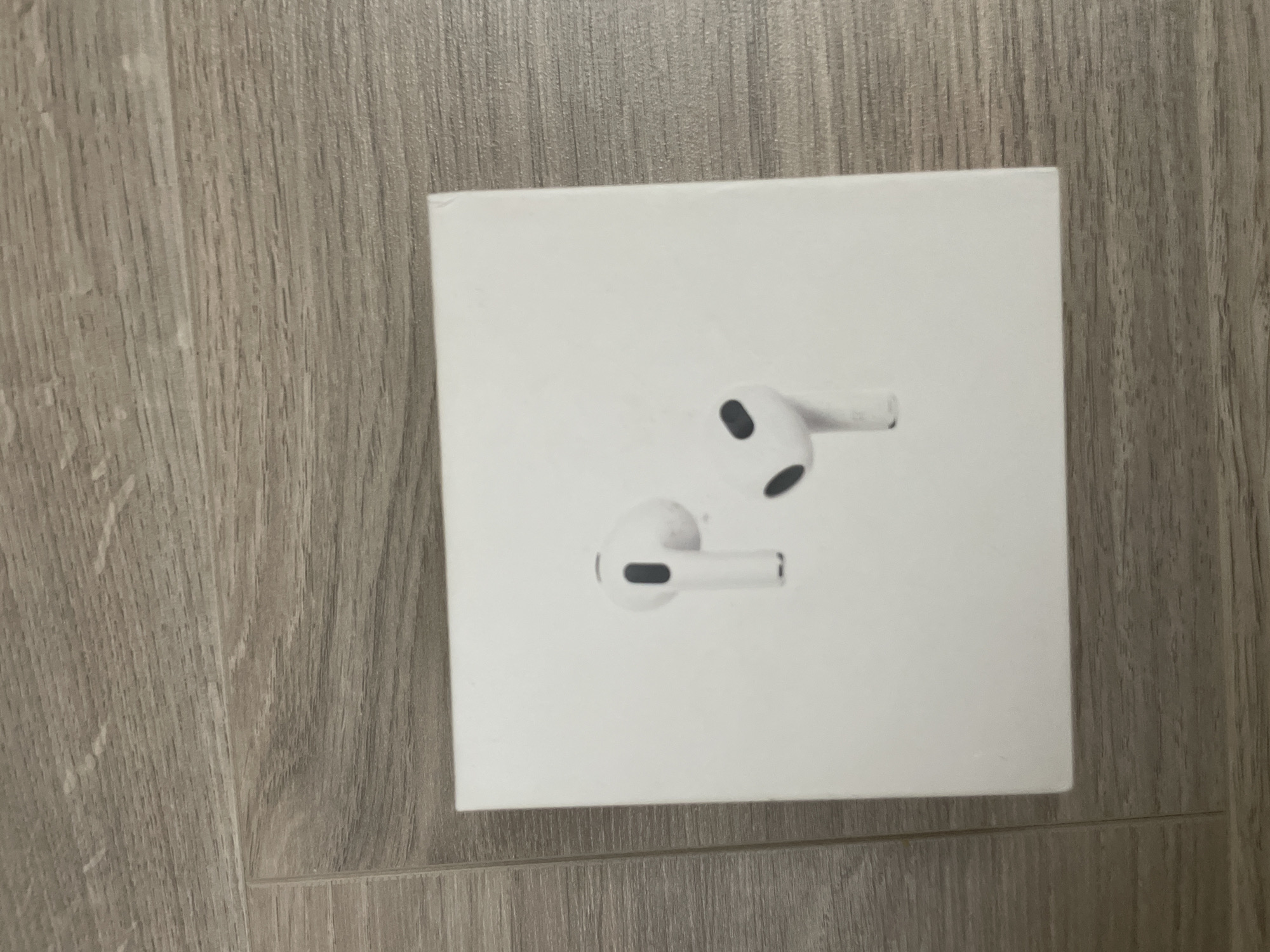 A white, rectangular product box is centrally positioned on a light-colored wooden surface. The box features a clear image of two white earbuds, resembling Apple AirPods. One earbud is depicted slightly above and to the left of the other, both angled downwards and to the right, showcasing black accents that are likely speaker grilles or sensors. The box itself is plain white and appears clean and new.
The wooden surface beneath the box exhibits prominent grain patterns, with the texture running vertically on the left side of the frame and horizontally along the bottom edge. The wood has a muted gray-brown tone, suggesting a natural or laminate finish. The lighting is soft and even, indicating an indoor setting without strong shadows or cues about the time of day.
No activity is taking place in the image; it is a static product shot. No explicit text or branding is visible on the box, though the design of the earbuds strongly implies the product type. The location context for this image is Ihtiman, Bulgaria.