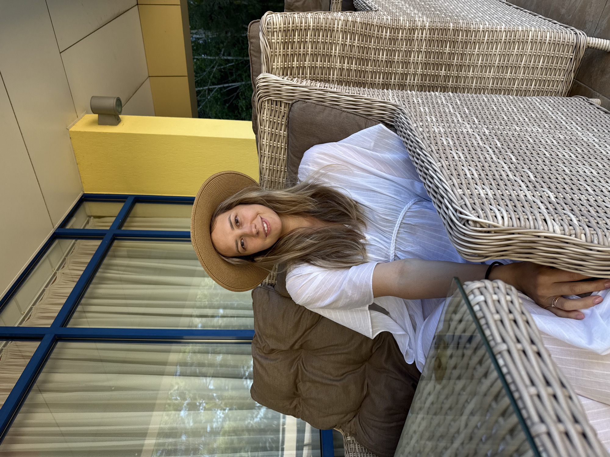 A young woman with blonde hair, wearing a wide-brimmed straw hat and a white dress, sits on a wicker chair on an outdoor patio. She is seated next to a glass-topped table and the patio has a yellow wall with a blue-framed window with sheer curtains. The background reveals trees and foliage. The woman is looking at the camera and smiling. She is wearing two rings on her left hand. This location appears to be an outdoor seating area of a hotel or restaurant in Nebug, Russia, judging by the architectural style and vegetation.