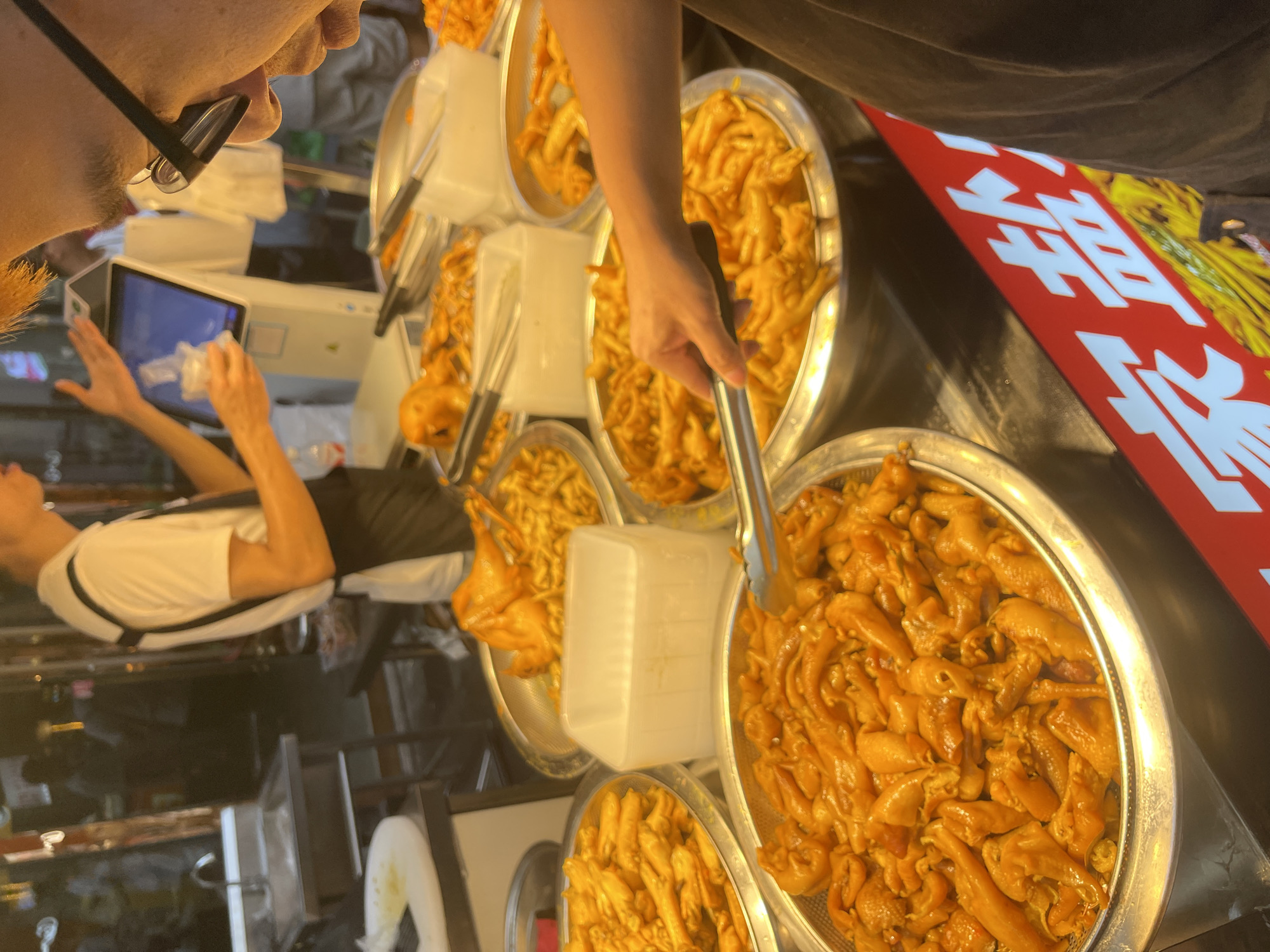The image shows a close-up, somewhat tilted view of a food stall in Panyu District, China. The primary subjects are several large, metal bowls overflowing with cooked, golden-brown food items that appear to be small intestines or similar internal organs, possibly prepared in a savory sauce. One bowl is in the foreground, with a hand holding metal tongs reaching into it.
In the background, a person wearing glasses is looking at a tablet device. Another person, wearing a white t-shirt, is leaning over a counter. The setting is a busy market or street food vendor, with other food items and containers visible, including a white plastic container and a colander filled with more of the food. A red sign with white Chinese characters is partially visible on the right side of the image, suggesting it's a point of sale or advertisement. The lighting suggests it could be daytime, possibly late afternoon or evening given the stall setting. The overall mood is one of commerce and food preparation. The visible text on the red sign is "雞蛋".