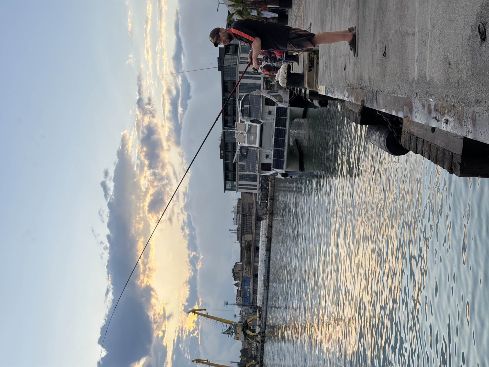 A man is depicted fishing from a concrete pier in a harbor or marina during the late afternoon or early evening, characterized by warm, golden light. He wears a dark t-shirt with orange trim, dark shorts, a baseball cap, and an orange chest strap or harness, holding a fishing rod extended over the water. A white container or cooler sits beside him on the pier.
Docked in the calm water is a prominent boat, likely a catamaran, featuring numerous solar panels installed on its roof and sides. In the background, across the water, an industrial landscape unfolds with large yellow cranes and various buildings, signaling an active port environment. The sky above is partly cloudy, with the setting sun casting golden and orange light on some clouds, while other parts of the sky remain a soft blue. The water's surface reflects the sky's light, creating shimmering patterns. The scene suggests a moment of peaceful recreation within the bustling context of a port in Burgas, Bulgaria. No legible text is visible in the image.