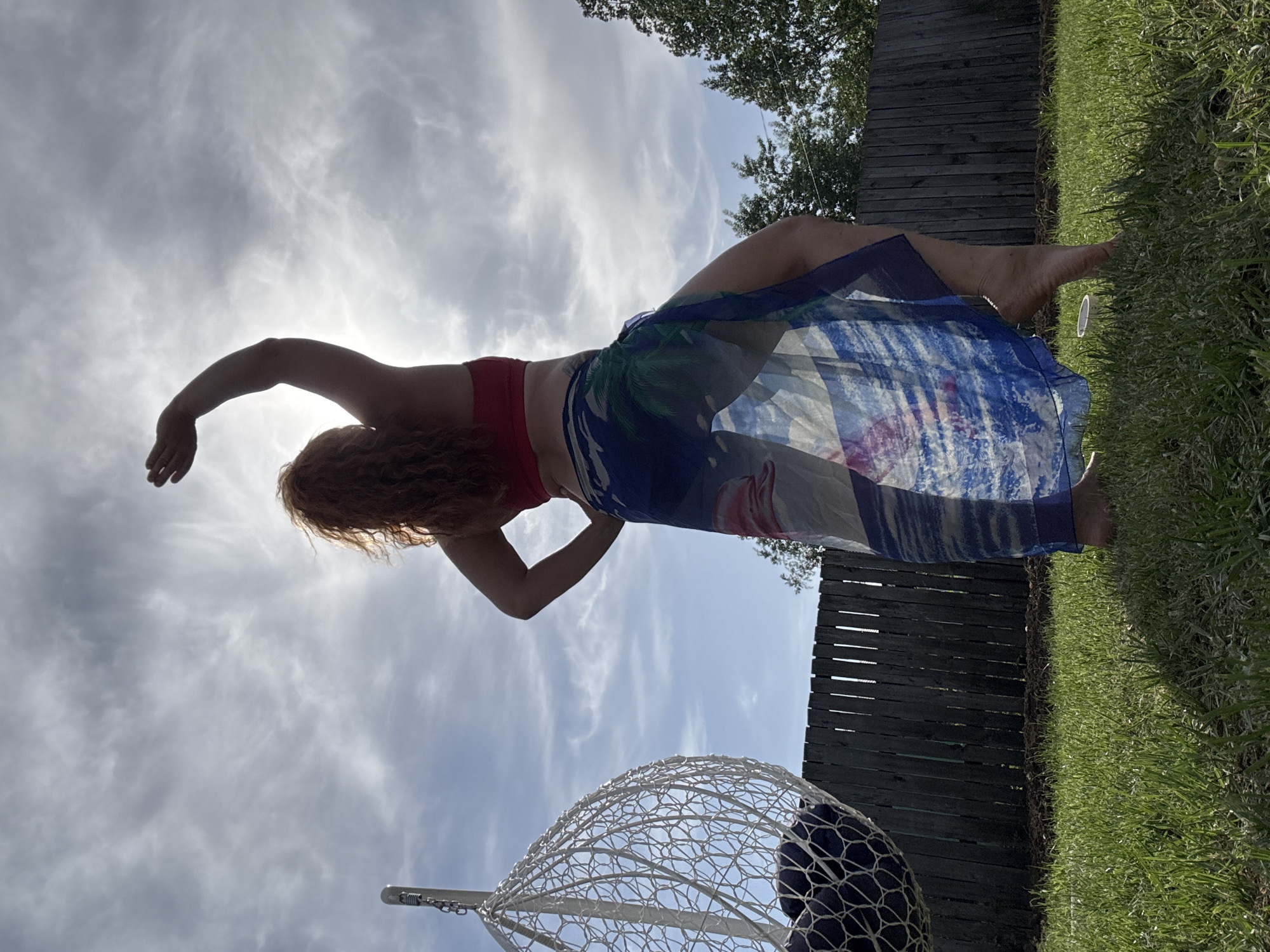 A woman with red hair is reclining outdoors, wearing a red top and a semi-transparent sarong with blue, green, and white patterns. She has one arm raised towards the sky, which is partly cloudy with the sun shining through. Her other arm is bent and placed behind her back. She is barefoot. A woven egg-shaped chair hangs to her lower left. The background includes a wooden fence, green grass, and some trees. There is no evidence suggesting this image was taken in Chaikovsky, Russia.