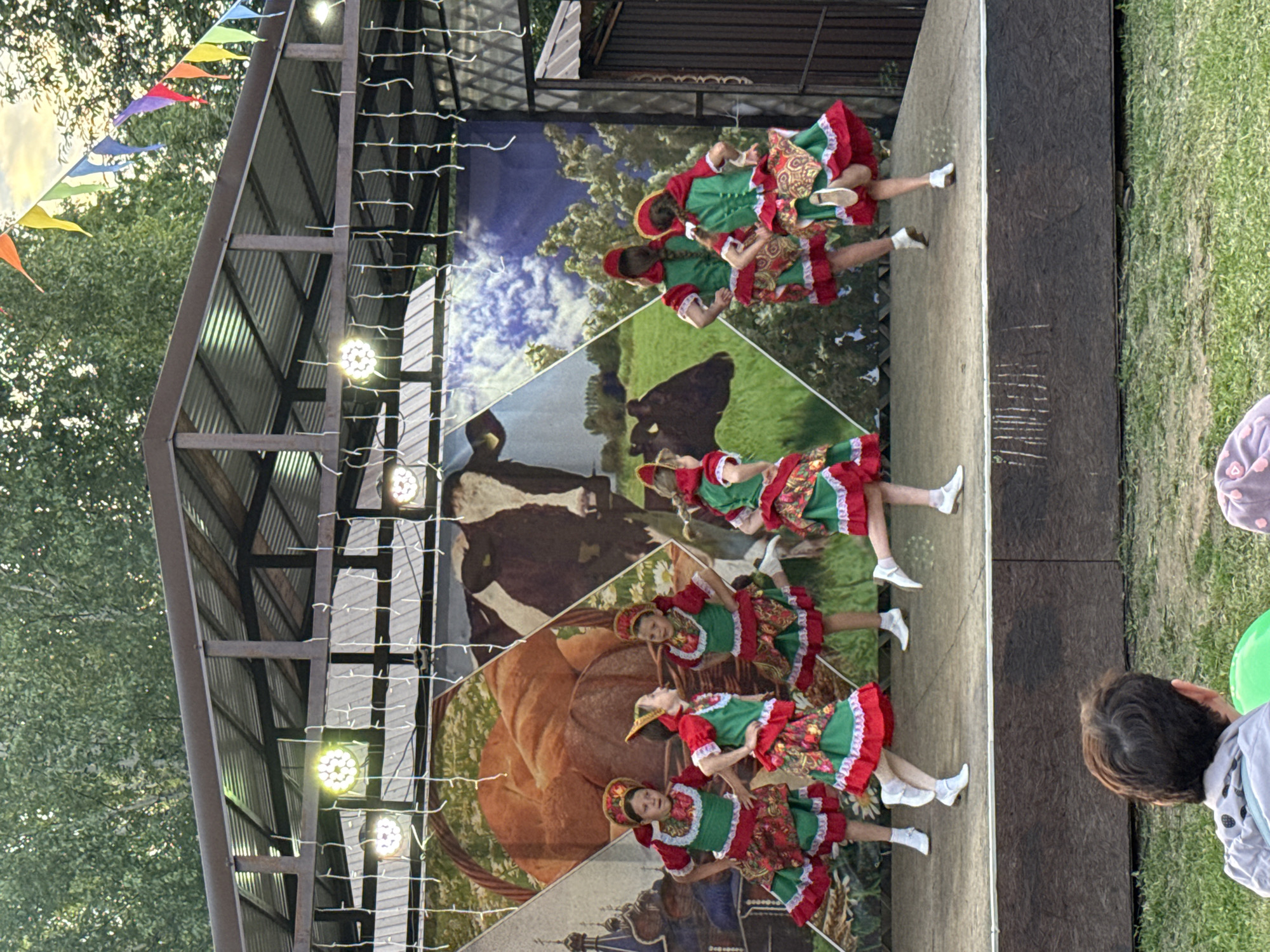 On an outdoor stage in the city of Kukui, Russia, a group of young girls in traditional Russian folk costumes are performing a dance. The costumes are predominantly green and red, with intricate patterns on the skirts and headdresses. The backdrop of the stage features a collage of images, including a large basket of bread, a cow in a grassy field, and what appears to be a Russian Orthodox church. Decorative lights and colorful triangular flags are strung across the top of the stage. The girls are in various poses, some mid-dance, showcasing their energetic performance. In the foreground, out of focus, are the backs of audience members seated on the grass, watching the performance. The setting appears to be a park or an open-air venue, with trees visible in the background.