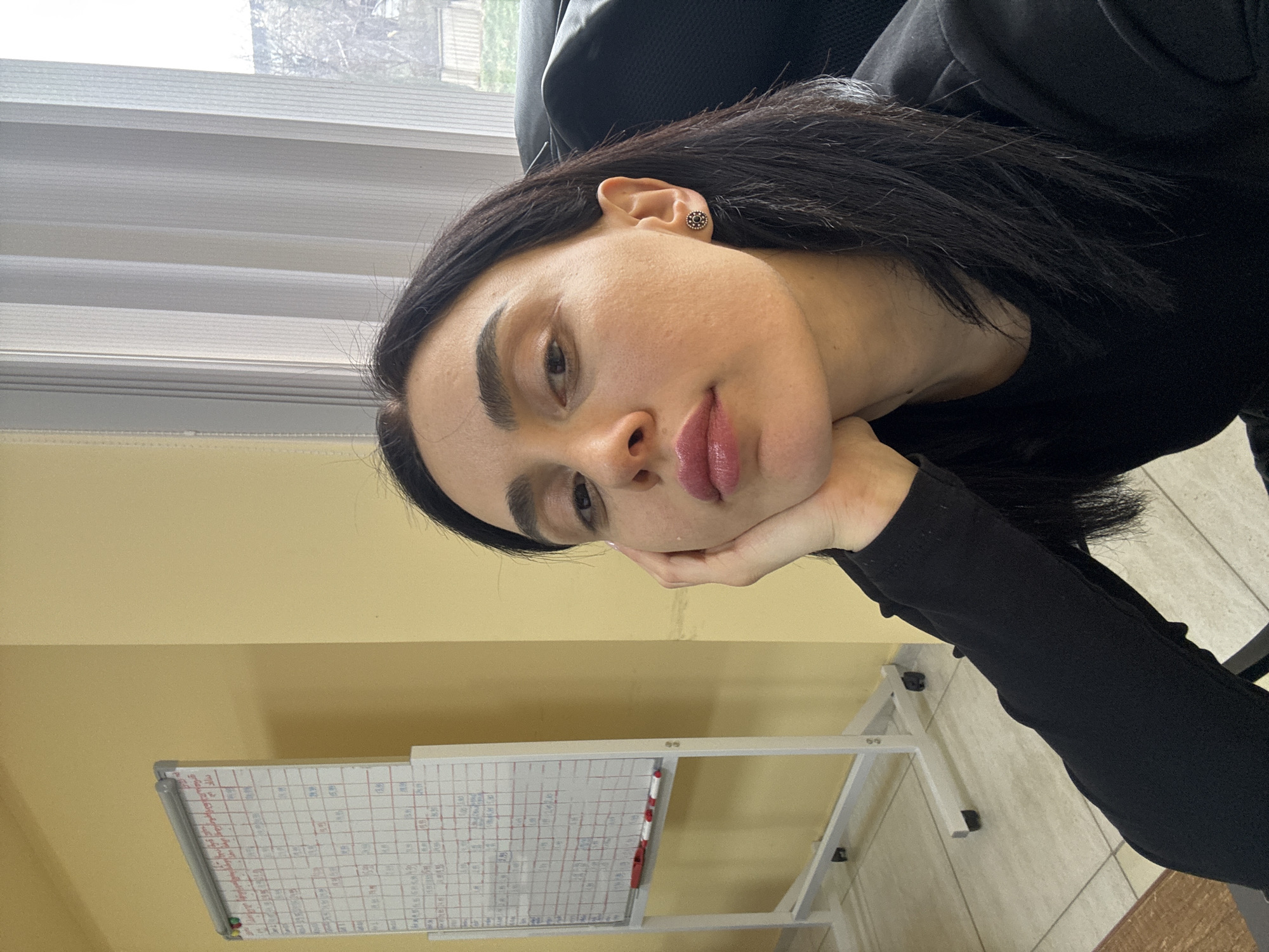 A woman with dark hair is resting her head on her hand. She is wearing a black top and an earring is visible in her left ear. Behind her is a window with horizontal blinds. A whiteboard with markings is positioned to the lower left of the frame. The setting is indoors, likely an office, with light yellow walls. Location in the city of Tbilisi, Georgia is unknown.