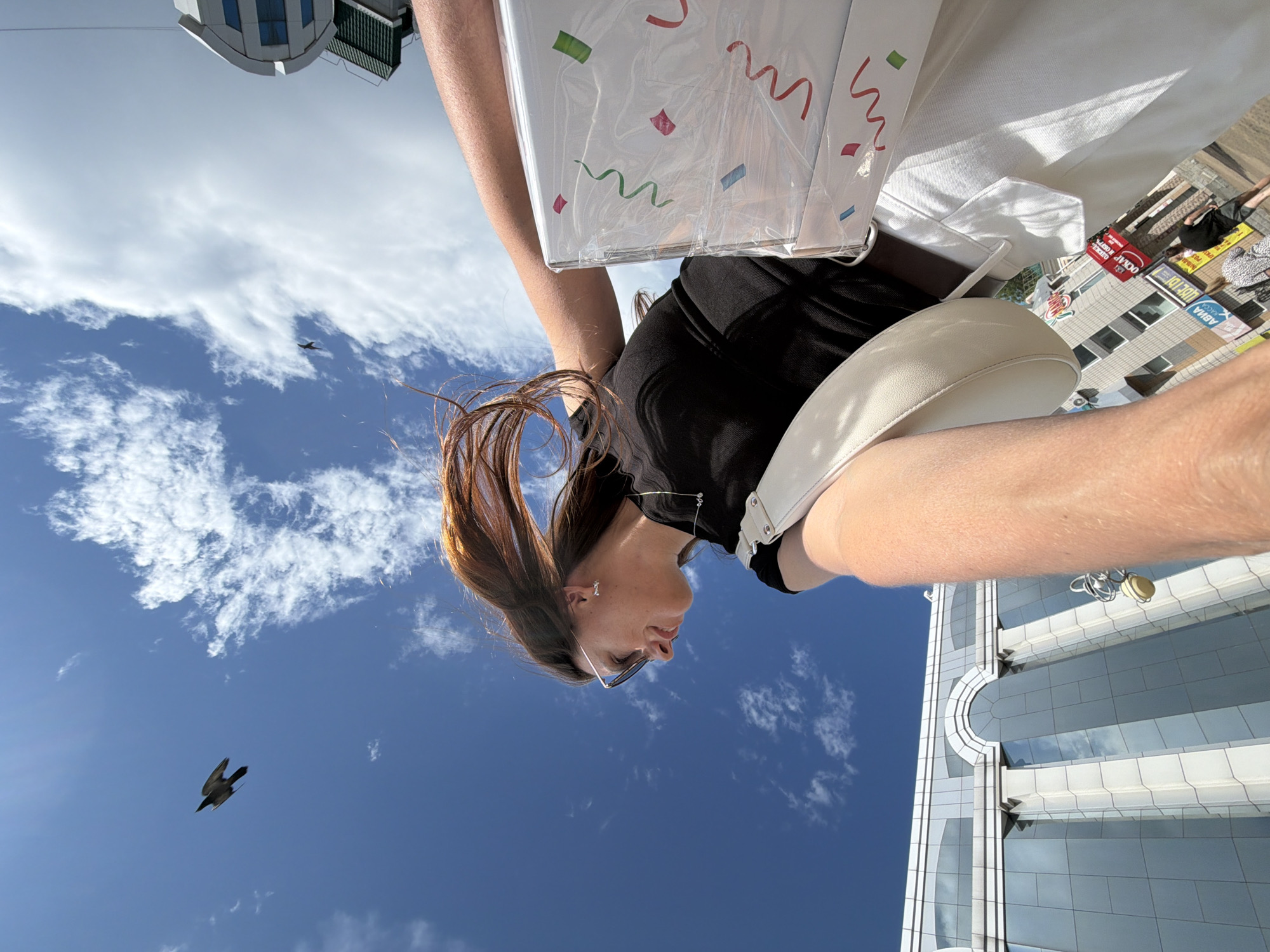 The image captures a low-angle perspective of a young woman with auburn hair, wearing sunglasses and a black top, as she walks outdoors. She is holding a white purse and carrying a white, rectangular package decorated with colorful confetti-like streamers. The background features a bright blue sky with scattered clouds and a bird in flight. To the left, a modern building with reflective glass windows is visible, indicative of an urban environment. The scene appears to be set in Birobidzhan, Russia, likely in a commercial or city center area, suggested by the presence of storefronts and people in the distance. The overall mood is casual and possibly festive, given the gift-wrapped package.