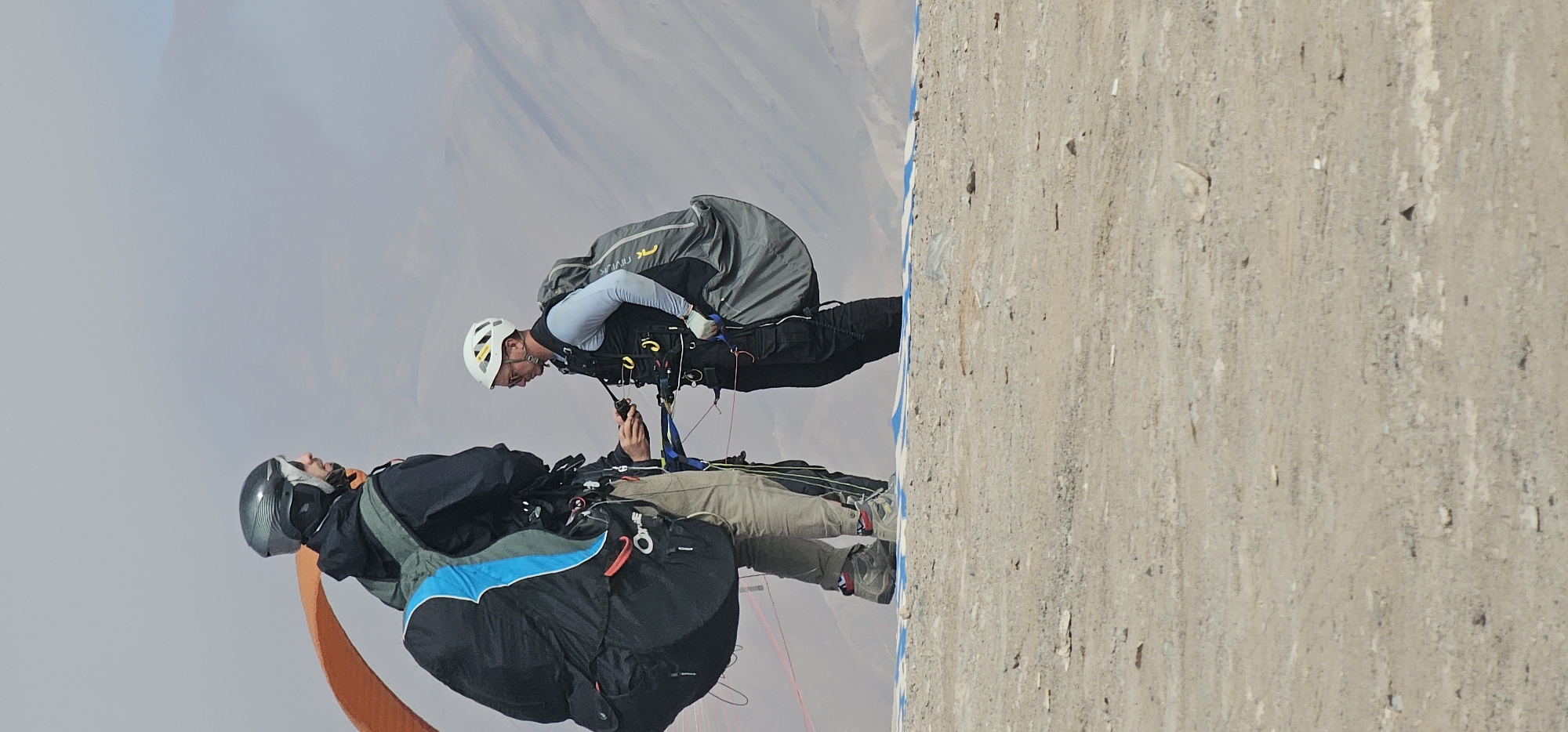 Two paragliders are mid-air near a concrete structure, presumably in preparation for landing or shortly after takeoff. One paraglider, wearing a dark helmet and black jacket with a blue accent, is in the foreground. The other, wearing a white helmet and grey long-sleeve shirt under a black vest, is slightly behind and to the right. They appear to be in tandem paragliding, possibly a passenger and pilot. The landscape in the background is arid, suggesting a desert or mountainous region. The location is Alto Hospicio, Chile.