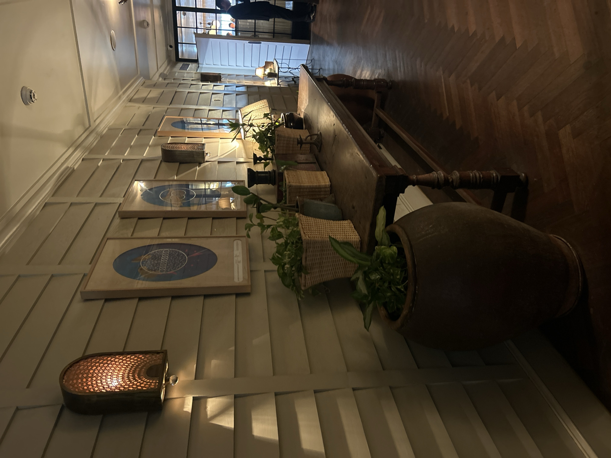 The image depicts a dimly lit interior hallway, likely in a hotel or restaurant, with a long wooden table as the central focus. The table is adorned with several potted plants in woven baskets and ceramic pots, as well as decorative items such as candle holders and a lamp. The walls are paneled with horizontal wooden planks and feature wall sconces that cast a warm glow. Framed circular artwork hangs on the wall behind the table. To the right, a doorway leads to another area where a person in a suit is visible standing. The floor is laid with dark wooden planks in a herringbone pattern. While the image does not explicitly state its location, the ambiance and decor suggest a sophisticated establishment, possibly a high-end hotel lobby or a restaurant in Mumbai, India, given the common architectural and interior design elements found in such establishments in the city.