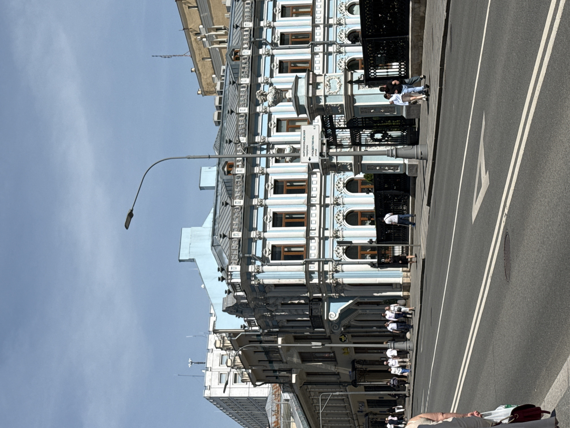 The image captures a street scene in Moscow, Russia. A light-blue and white, ornate building dominates the left side of the frame, its facade featuring multiple windows and intricate architectural details. A street sign, partially obscured, indicates "Варсонофьевский" (Varsonofyevskiy) and "Рождественка" (Rozhdestvenka). Adjacent to the building is a sidewalk with people strolling along it. The right side of the image depicts a paved road with double white lines dividing traffic lanes. A manhole cover is visible on the road surface. The sky above is clear and light blue. The foreground shows part of a person and personal belongings, suggesting the photo was taken from a low vantage point on the ground.