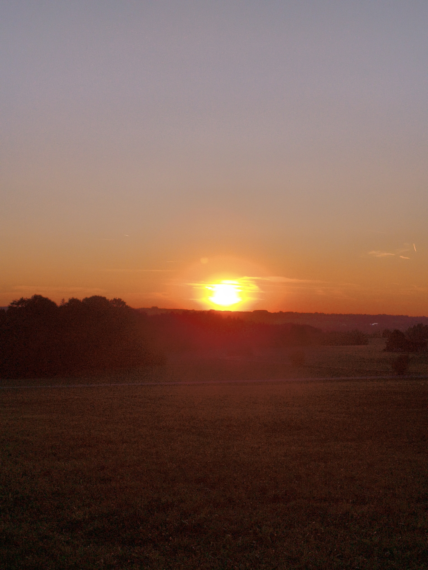 This is a photograph of a sunset over a rural landscape. The sun, a bright yellow orb, is low on the horizon, casting a warm orange glow across the sky. The sky transitions from a pale purple at the top to a vibrant orange near the horizon. In the foreground, there is a dark silhouette of trees and rolling hills. Beyond the hills, the land appears to be a field or meadow, also cast in the warm light of the setting sun. The overall impression is one of peace and natural beauty. The photograph was taken in Mauerstetten, Germany.