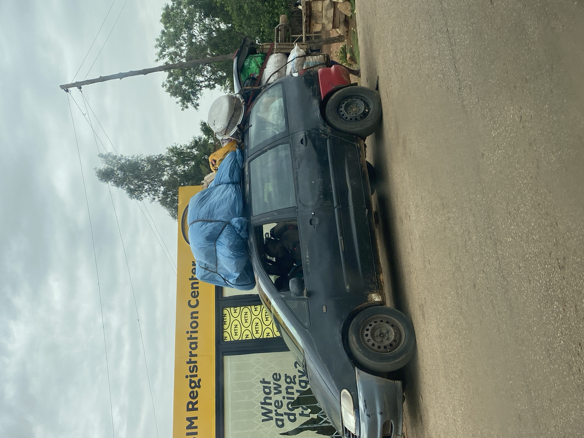 The image shows a black car heavily loaded with various items on its roof. The car is parked on a paved road in front of a building with a yellow sign that reads "SIM Registration Center" with "MTN" logos repeated in the windows. The text "What are we doing today?" is visible on the window below the logos. Above the car, utility poles with power lines stretch across the sky. Trees are present in the background. The overall impression is that the car is overloaded, potentially exceeding safety limits.