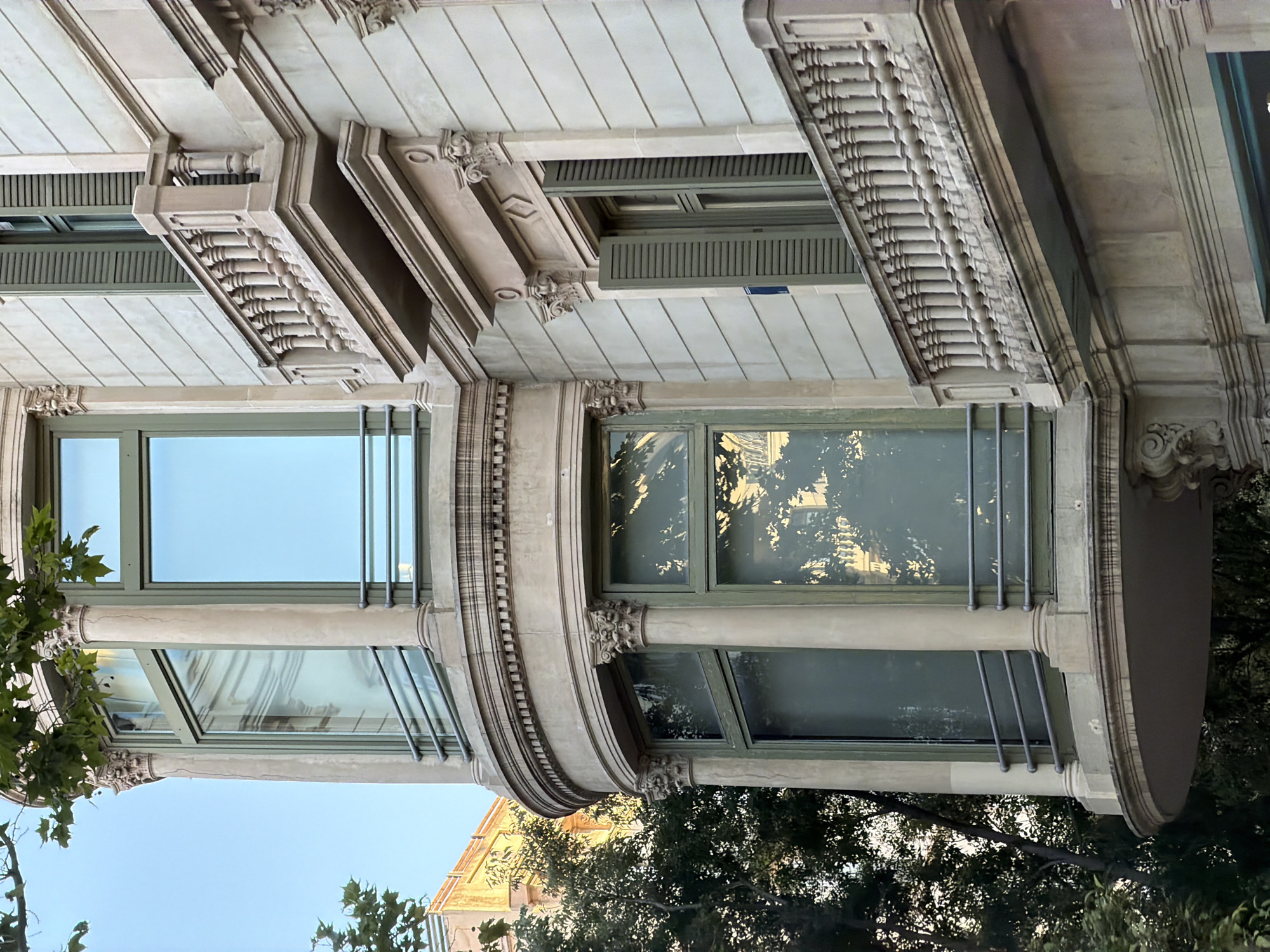 The image shows the exterior of a building in Barcelona, Spain. The architecture features a curved bay window with large glass panes framed in a light green color. The window is supported by Corinthian columns. Adjacent to the bay window is a balcony with a balustrade made of stone spindles. Another window with green shutters is visible on the right side of the building. The building's facade is made of light-colored stone, with decorative moldings and carvings. Trees with green foliage are visible in the foreground and to the left of the building. The sky in the background is clear and blue. The scene is captured from a slightly low angle, looking up at the building.