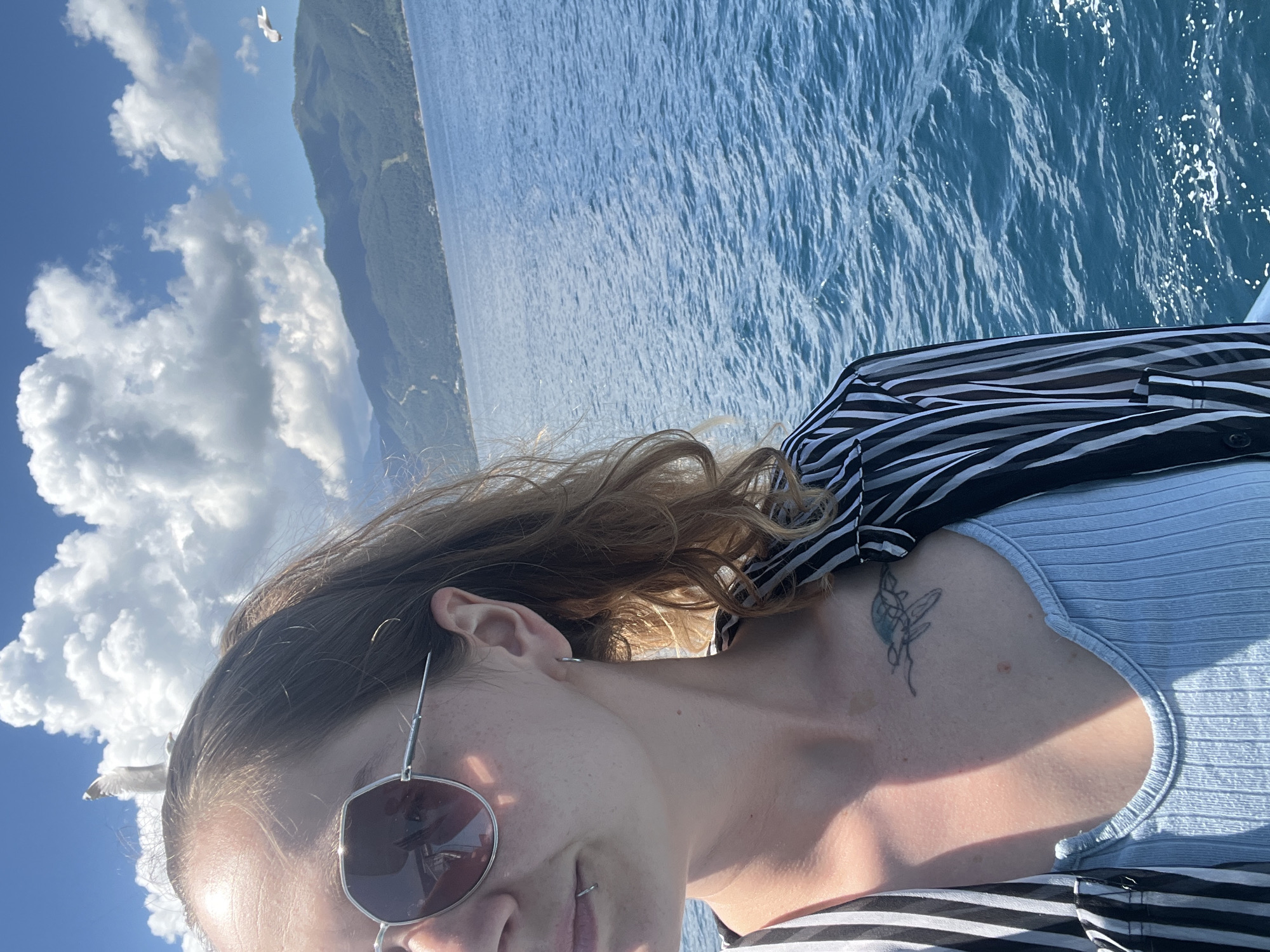A woman is captured in a close-up selfie with a backdrop of the sea and mountains. She is wearing sunglasses and a striped shirt over a light blue top. A tattoo of a sea turtle is visible on her chest. The sea is a vibrant blue, with small waves. In the distance, a mountainous coastline is visible under a partly cloudy sky. Two seagulls are present, one perched on the woman's head and another flying in the distance. The image appears to be taken on a boat during the day. The presence of the sea and mountainous coastline suggests a location like Thassos, Greece, known for its natural beauty and proximity to the sea.