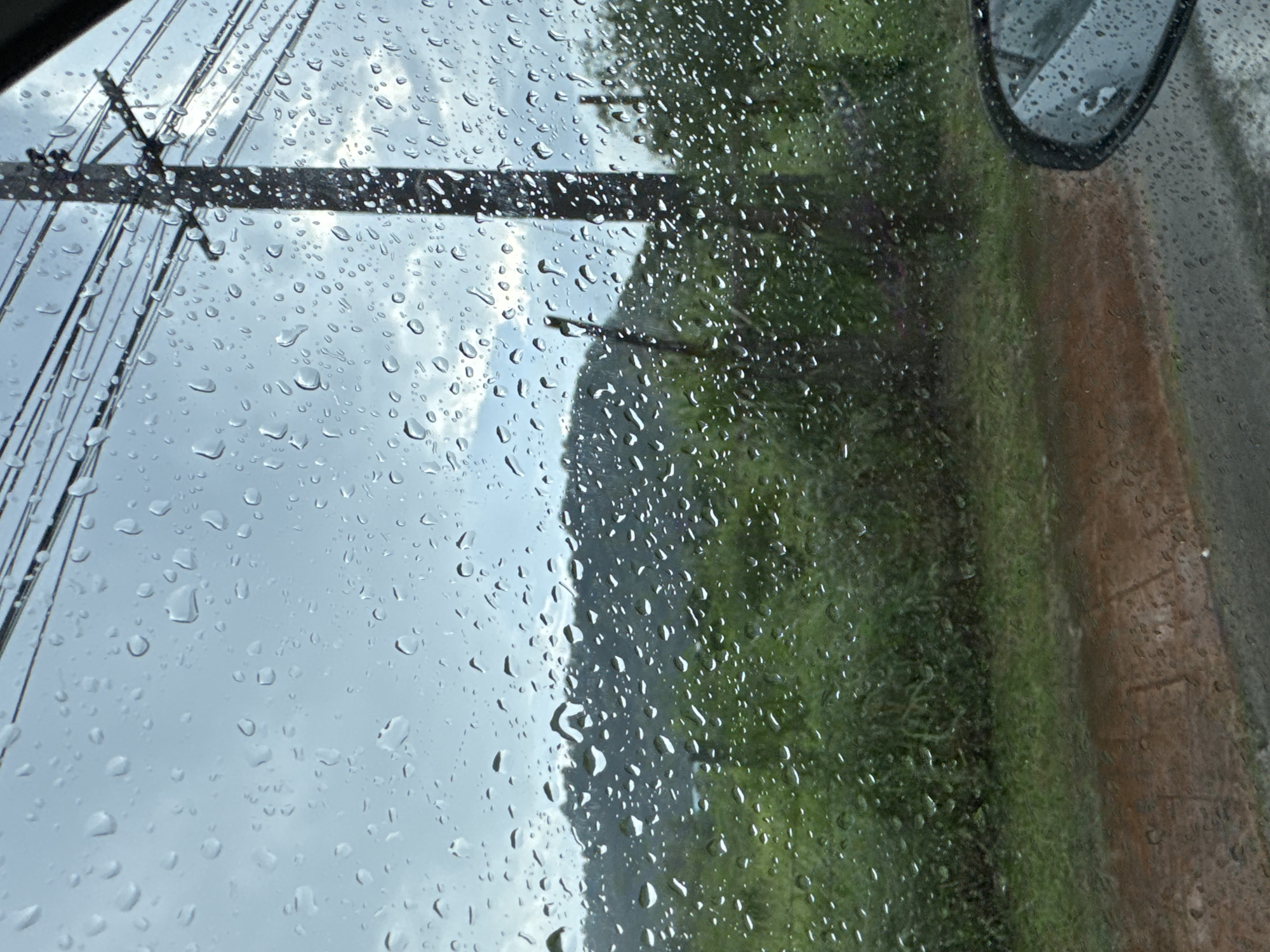 The image depicts a view through a rain-streaked window, likely from a vehicle, looking out at a cloudy, overcast sky. In the mid-ground, a dense area of green vegetation, possibly grass or shrubs, is visible, with a sloping hill or mountain range in the background, also covered in greenery. A utility pole with multiple wires extends vertically on the right side of the frame. The foreground shows a wet, reddish-brown surface, possibly a dirt road, and a portion of a car's side mirror is visible on the bottom right. The environment suggests a rural or semi-rural setting, possibly during or after rainfall. No people or specific actions are clearly discernible.