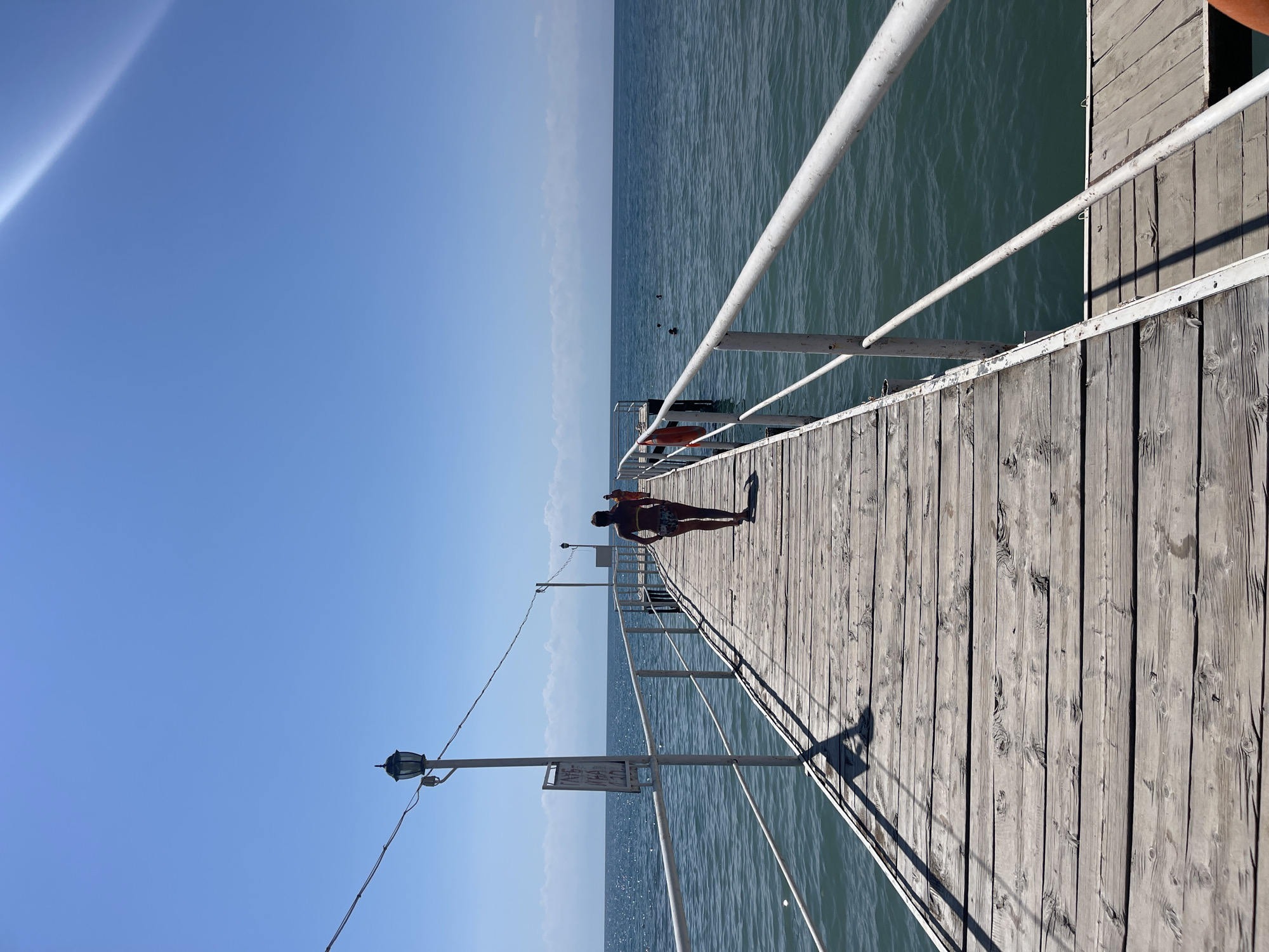 A lone woman walks away from the camera down a weathered wooden pier that extends into a calm, blue body of water under a clear sky. The pier is bordered by a white railing, and a lamppost with a sign is visible on the left. In the distance, several other people can be seen swimming in the water. The scene appears to be a resort or recreational area.