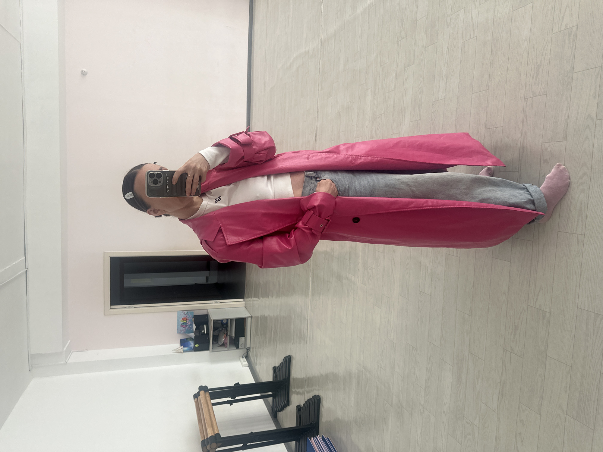 A person in a pink coat takes a selfie in a room. The room has light-colored wood flooring, white walls, and a shelf with items. There is also exercise equipment present. The location is likely indoors, but the city of Votkinsk, Russia, is not ascertainable from the image.