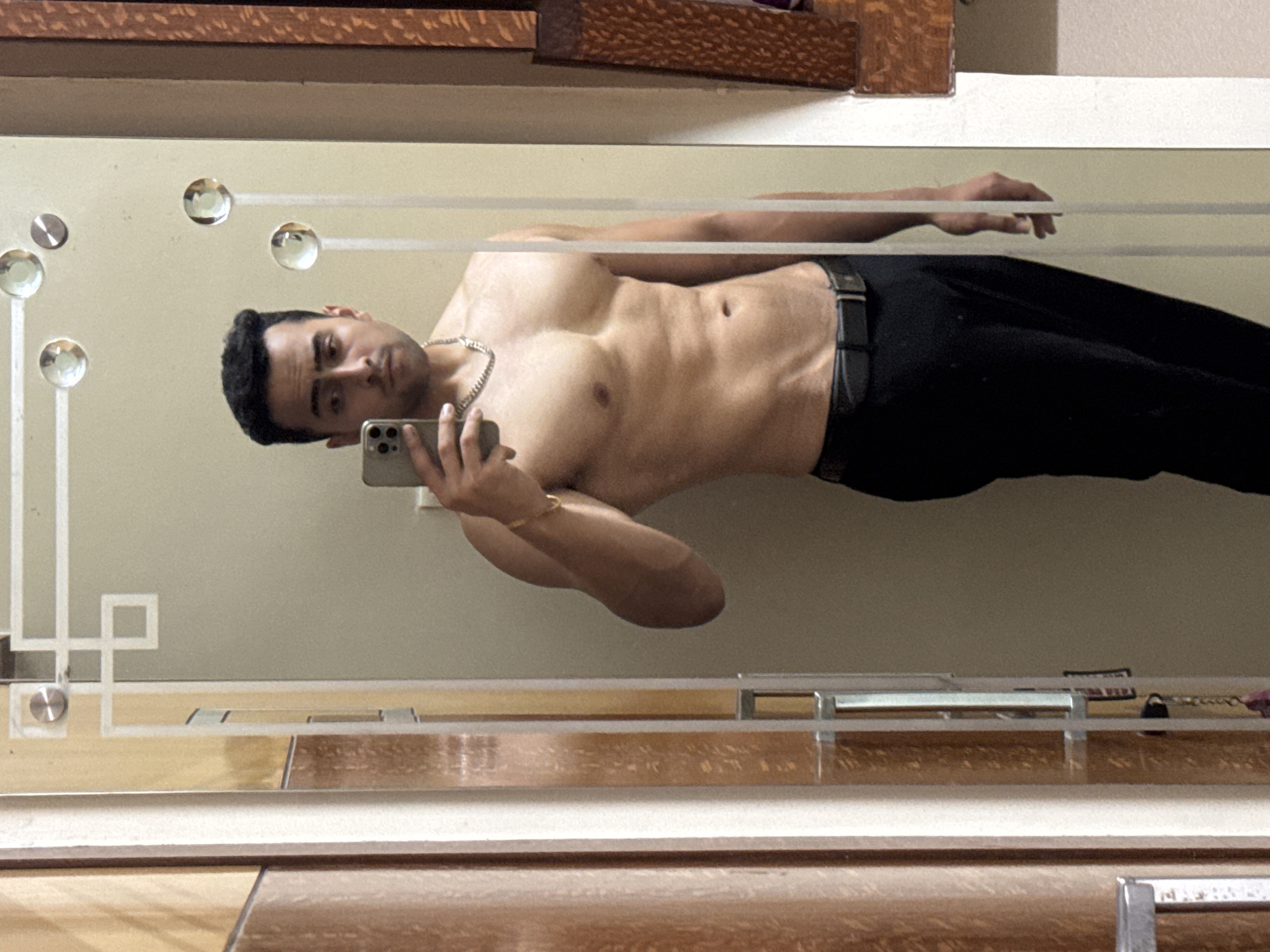 A man is taking a shirtless selfie in a mirror. He is wearing black pants and a silver chain necklace. He is holding a phone in his left hand. The mirror is framed with a decorative design and is mounted on a wooden dresser. The location is unknown; there is no indication it is in Kolkata, India.