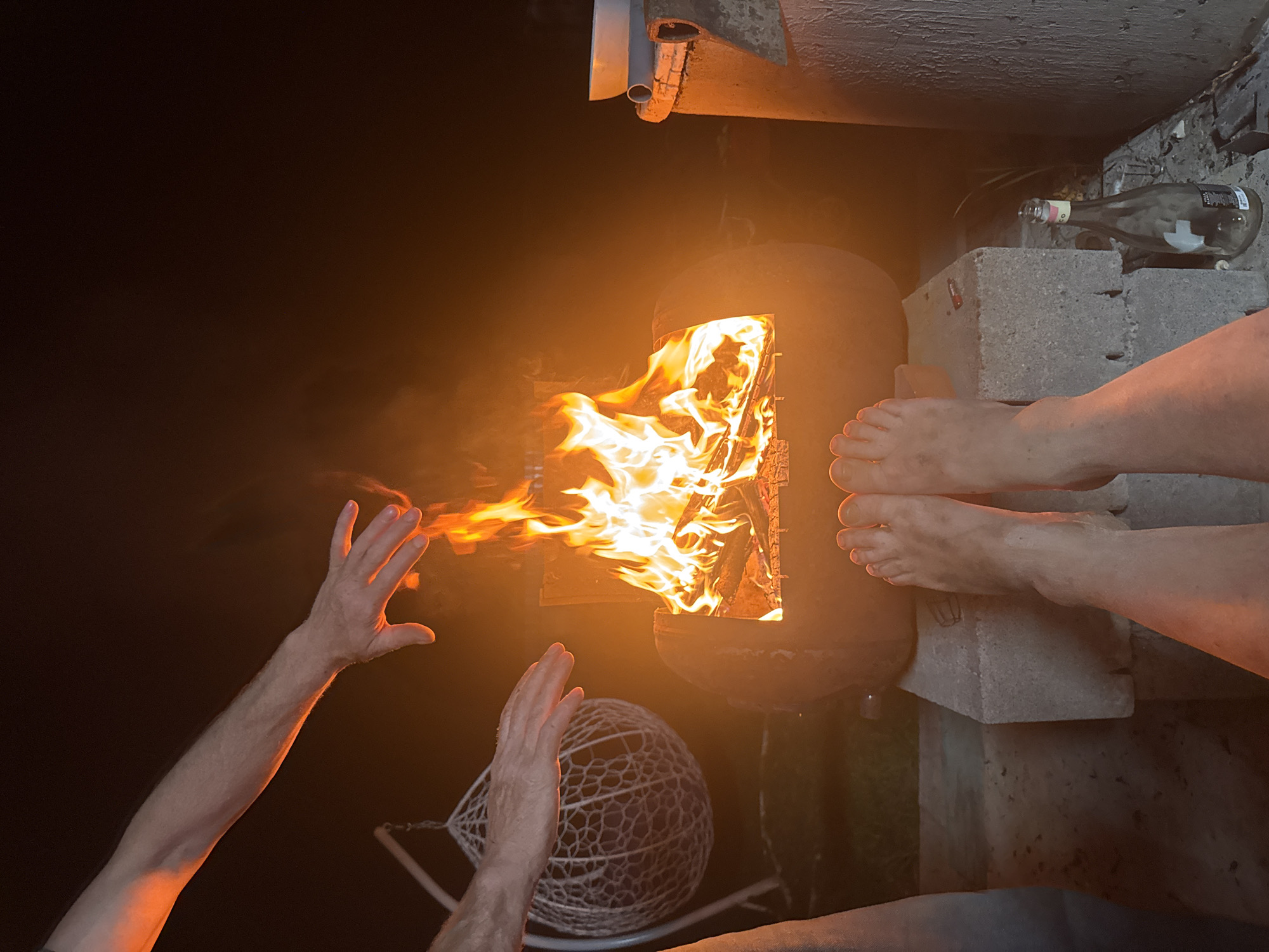A nighttime scene illuminated by a wood-burning stove containing a roaring fire. A person's hands are outstretched towards the flames as if warming them. Another person's bare feet are visible resting on concrete blocks near an empty wine bottle. A webbed lawn chair is nearby. The location is Chaikovsky, Russia.