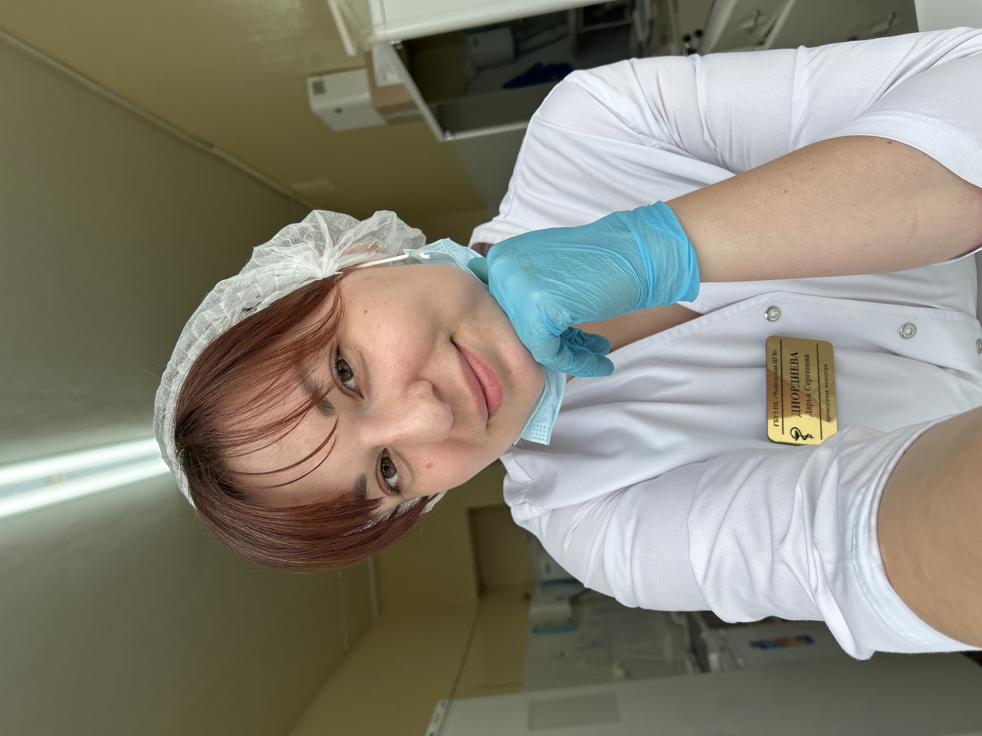 A young woman is featured in a selfie, taken from a slightly tilted, low-angle perspective, likely in a medical or clinical setting in Фоки, Russia. She has reddish-brown hair, dark eyes, and a pleasant, gentle smile, looking directly at the camera.
She is dressed in a white medical uniform coat over a white garment, with a white disposable hairnet covering her hair. A blue disposable medical glove is worn on her right hand, with which she is holding down a light blue disposable surgical mask, pulled below her chin to reveal her face.
The background consists of light-colored walls, indicative of an indoor institutional environment. Hints from the cropped images suggest the presence of fluorescent lighting and possibly some medical equipment or cabinetry.
A golden name tag is pinned to the right side of her white coat. The text on the badge is in Russian and identifies her:
"ГБУ ПК "Чайковского ЦРБ"" (State Budgetary Institution of Perm Krai "Chaykovsky Central District Hospital")
"ДИОРДИЕВА" (Diordieva - Last Name)
"Дарья Сергеевна" (Darya Sergeevna - First Name and Patronymic)
"процедурная медсестра" (procedural nurse / treatment nurse).
The image captures a moment of a healthcare professional during her workday.