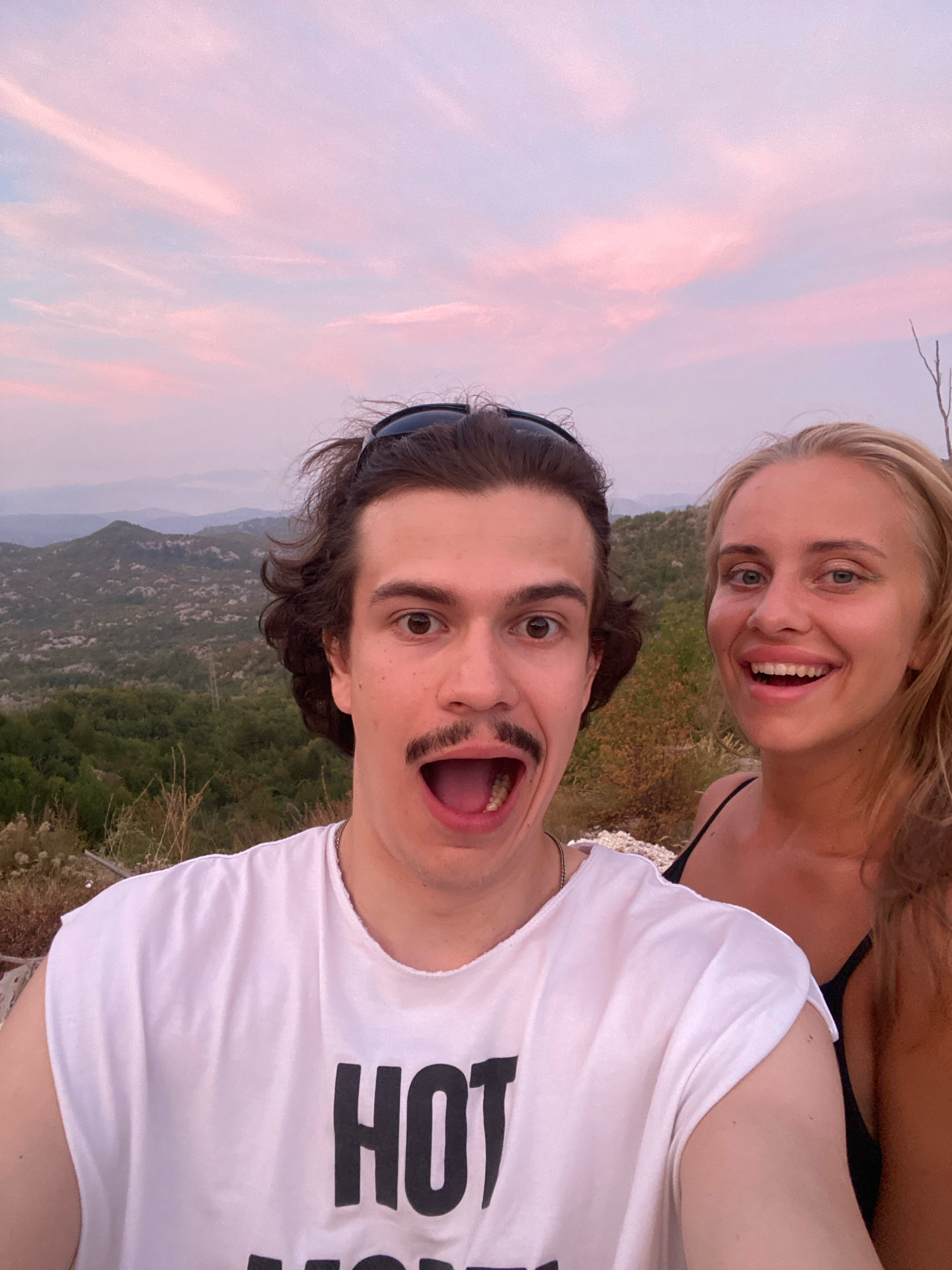 The image is a selfie taken outdoors with a scenic background of rolling hills and a pink-hued sky at dusk. In the foreground, a young man with dark, curly hair and a prominent mustache is wearing a white, sleeveless t-shirt with the word "HOT" printed in large black letters. He has sunglasses perched on his head and an amused expression with his mouth open. To his right, a young woman with blonde hair is smiling broadly, her mouth also open. She is wearing a black top. The background consists of lush green vegetation covering the hills, with some dry brush in the immediate foreground. The sky is filled with soft clouds reflecting shades of pink and purple, indicative of sunset. The overall impression is one of a joyful, candid moment captured during an outing. The location is identified as Boguti, Montenegro, suggesting a natural, possibly mountainous, environment.