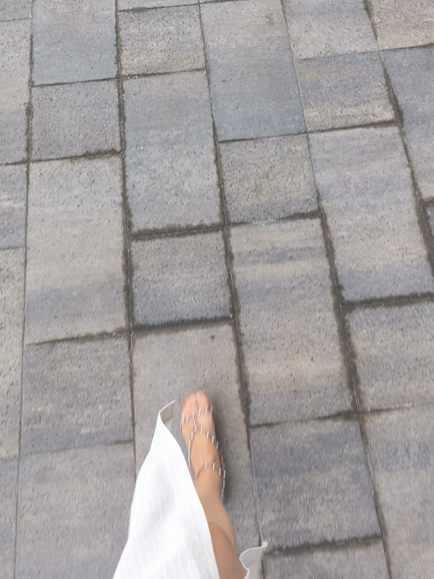 The image shows a person's foot wearing a sandal with thin straps adorned with small, reflective elements, walking on a paved walkway composed of large, rectangular gray stone tiles. A portion of a white garment, possibly a dress or skirt, is visible in the lower left of the frame. The walkway appears slightly wet in places, with darker patches indicating moisture. The scene is generic and lacks any specific visual cues that would indicate its location within the city of Novosibirsk, Russia, or any other specific urban environment. The focus is on the foot and the immediate paving stones.