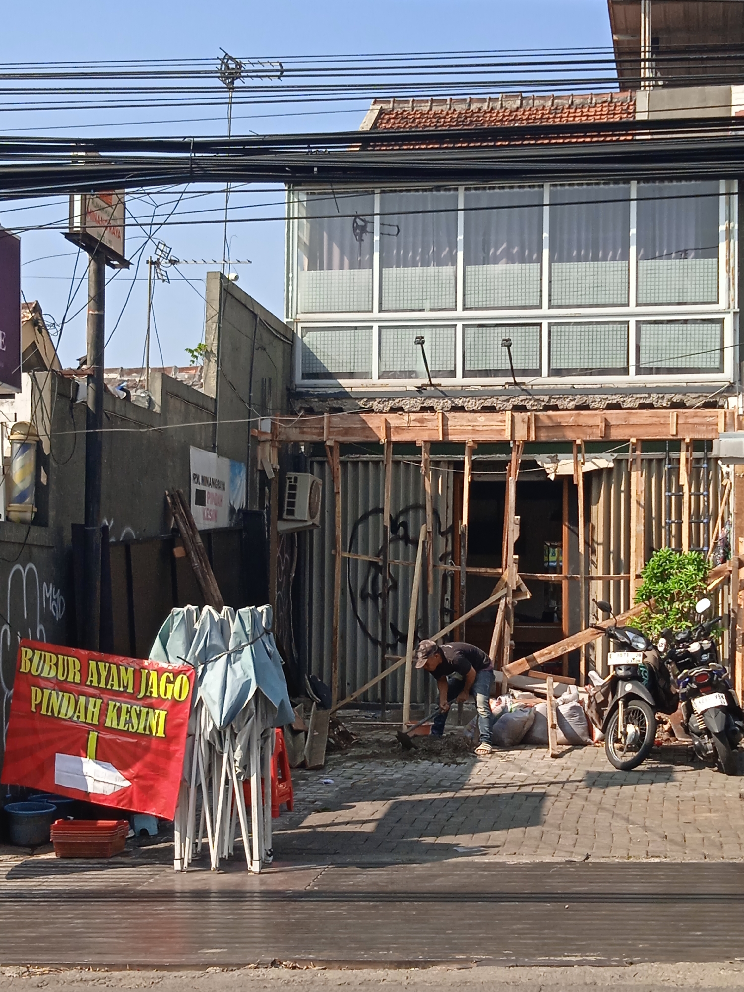 The image shows a street scene in Bandung City, Indonesia, with a building undergoing renovation. A person is digging in the foreground, and two motorcycles are parked nearby. A red banner with yellow text reads "BUBUR AYAM JAGO PINDAH KESINI," which translates to "Chicken Porridge JAGO Moved Here." The building's facade is partially dismantled, with wooden scaffolding and corrugated metal sheeting visible. Power lines and an antenna are present above the building. The sky is clear and blue.