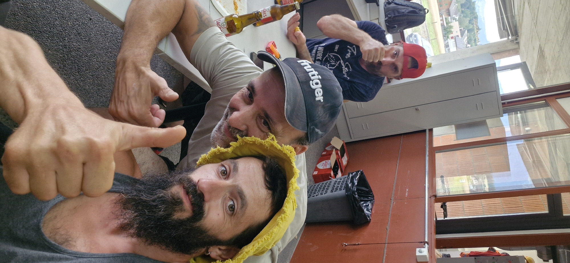 The image shows two men in an indoor setting, possibly an office or workshop. One man is wearing a grey t-shirt and a black baseball cap with "huttiger" written on it, and he is giving a thumbs-up gesture. The other man is wearing a blue t-shirt and a red baseball cap, and he is also giving a thumbs-up gesture. Both men appear to be happy and relaxed.
In the background, there are several objects, including a table with bottles of beer, a black backpack, a grey locker, and a trash can. The overall impression is that of a casual gathering or break time. The lighting suggests it is daytime. The image also includes a partial view of the exterior through a window, showing buildings and trees, hinting at an outdoor environment beyond the immediate setting.
There is text visible on the beer bottles and on the t-shirt of the man in the red cap. The t-shirt has white text that reads "Since 1919", "Work", and "Start". The bottles have text that appears to be in Portuguese.