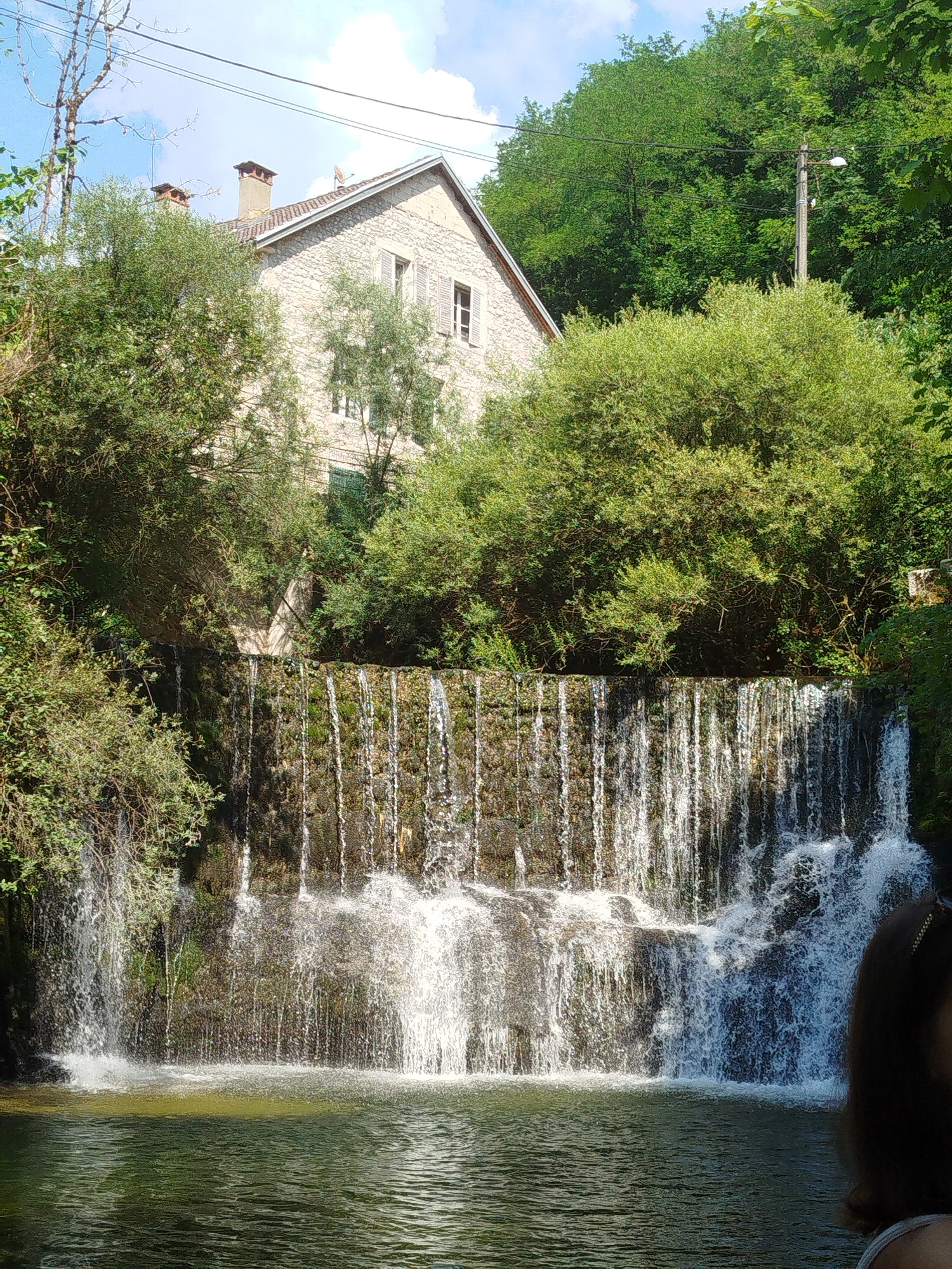 This image depicts a picturesque waterfall in a natural setting, likely in Saint-Claude, France. A stone building, possibly a house or an old mill, is visible in the background, partially obscured by lush green trees. The building has a pitched roof with chimneys and windows. The waterfall cascades over a rugged, moss-covered stone wall into a pool of tranquil, green water. The scene is illuminated by bright sunlight, suggesting it was taken during the daytime. The presence of trees and the natural flow of water indicate a rural or semi-rural environment. In the foreground, the edge of a person's dark hair and shoulder can be seen on the right side of the frame, suggesting someone is present and observing the waterfall. The overall mood is serene and idyllic, evoking a sense of natural beauty and tranquility. There is no visible text in the image.