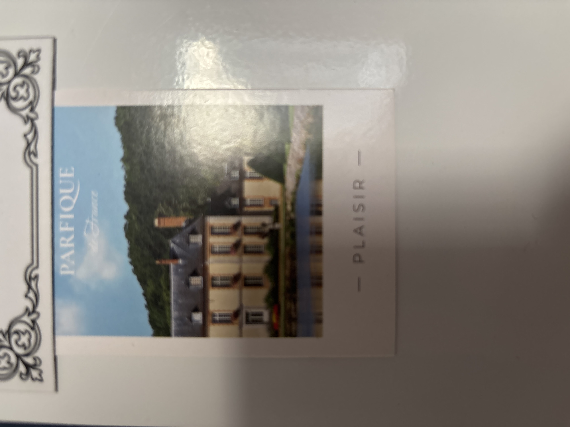 The image shows a postcard with a photograph of a French chateau or mansion. The chateau is a light yellow building with a dark grey roof and dormer windows. It is surrounded by trees and a body of water that reflects the building. Above the chateau, a blue sky with some clouds is visible. Text on the postcard reads "PARFIQUE de France" at the top, and "- PLAISIR -" at the bottom. The postcard is partially obscured by a white border and appears to be placed on a white surface. The overall impression is that of a travel souvenir or promotional item. There is no information in the image or its context to suggest any connection to the city of Imeni Morozova, Russia.