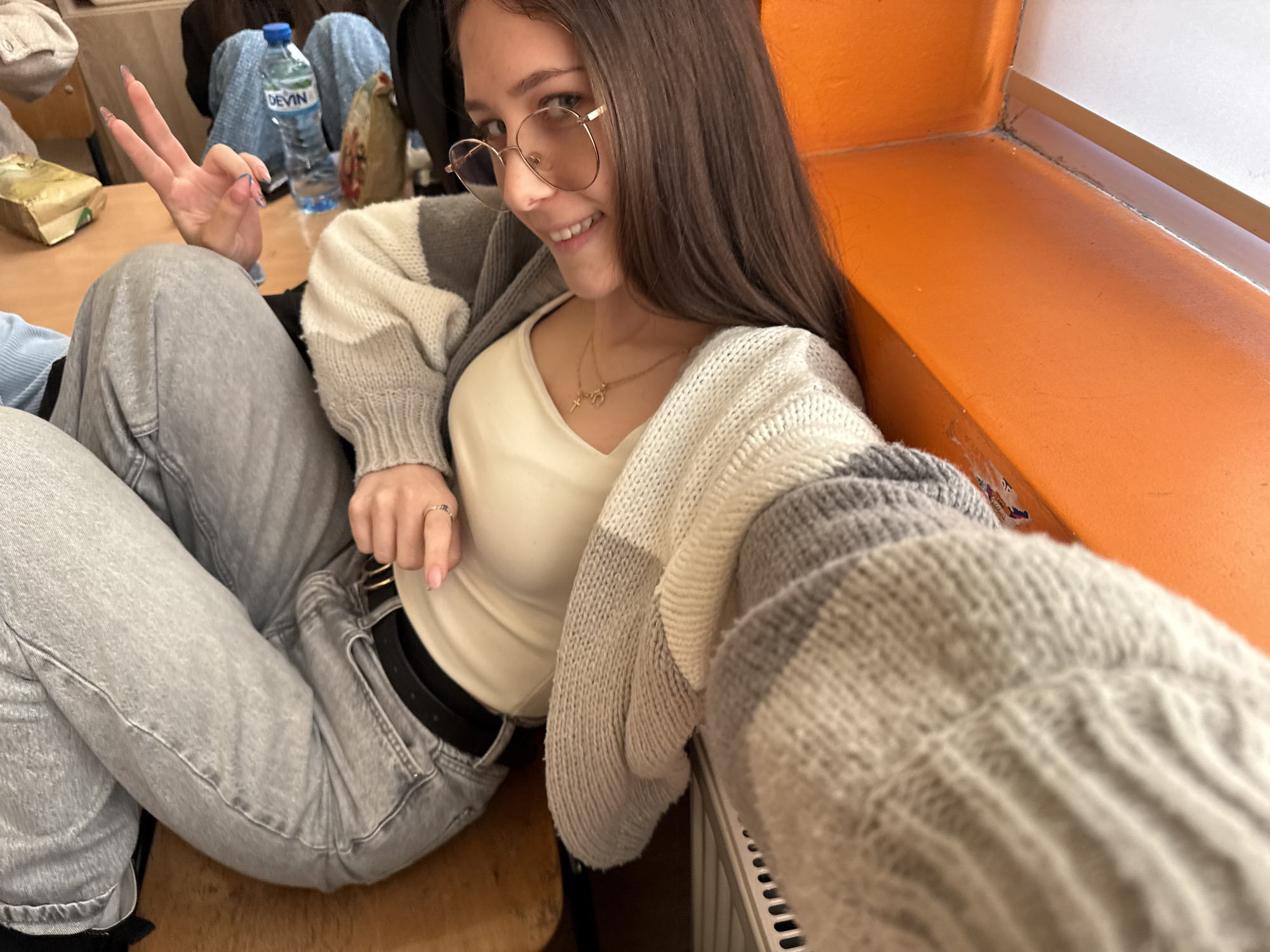 A young woman with long brown hair and round, gold-rimmed glasses smiles directly at the camera, making a peace sign with her left hand. She is dressed in a light cream-colored V-neck top, a grey, white, and black striped knit cardigan, and light grey jeans secured with a black belt. A delicate golden necklace with two small charms hangs around her neck, and a ring is visible on her right hand. Her fingernails are long and painted.
She is seated indoors, likely in a casual setting such as a classroom or common area, with what appears to be a light brown wooden surface beside her. In the background, a plastic bottle of "DEVIN" mineral water is visible on the surface, along with a wrapped package. The room features a distinctive bright orange wall or window sill that occupies the right side of the frame, with a sliver of a white window frame visible above it. Part of a radiator can be seen in the bottom right corner. The overall mood is casual and friendly, with the woman appearing cheerful. The presence of the "DEVIN" water bottle strongly suggests the location is Sofia, Bulgaria.
